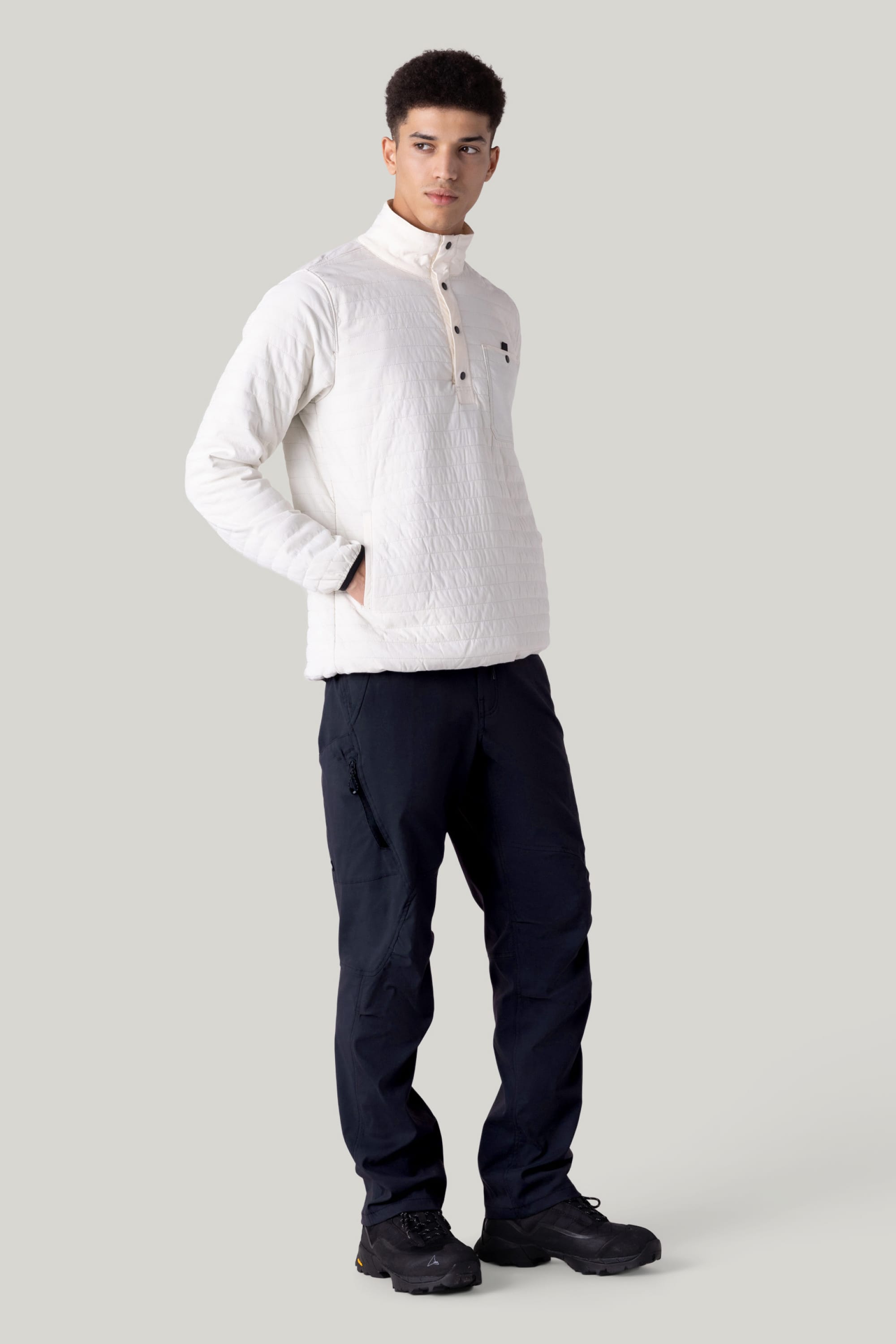 A man stands with hands in his pockets, wearing a white quilted pullover, 686 Men’s Anything Cargo Pant - Relaxed Fit by 686 in navy, and black shoes against a plain light gray background.