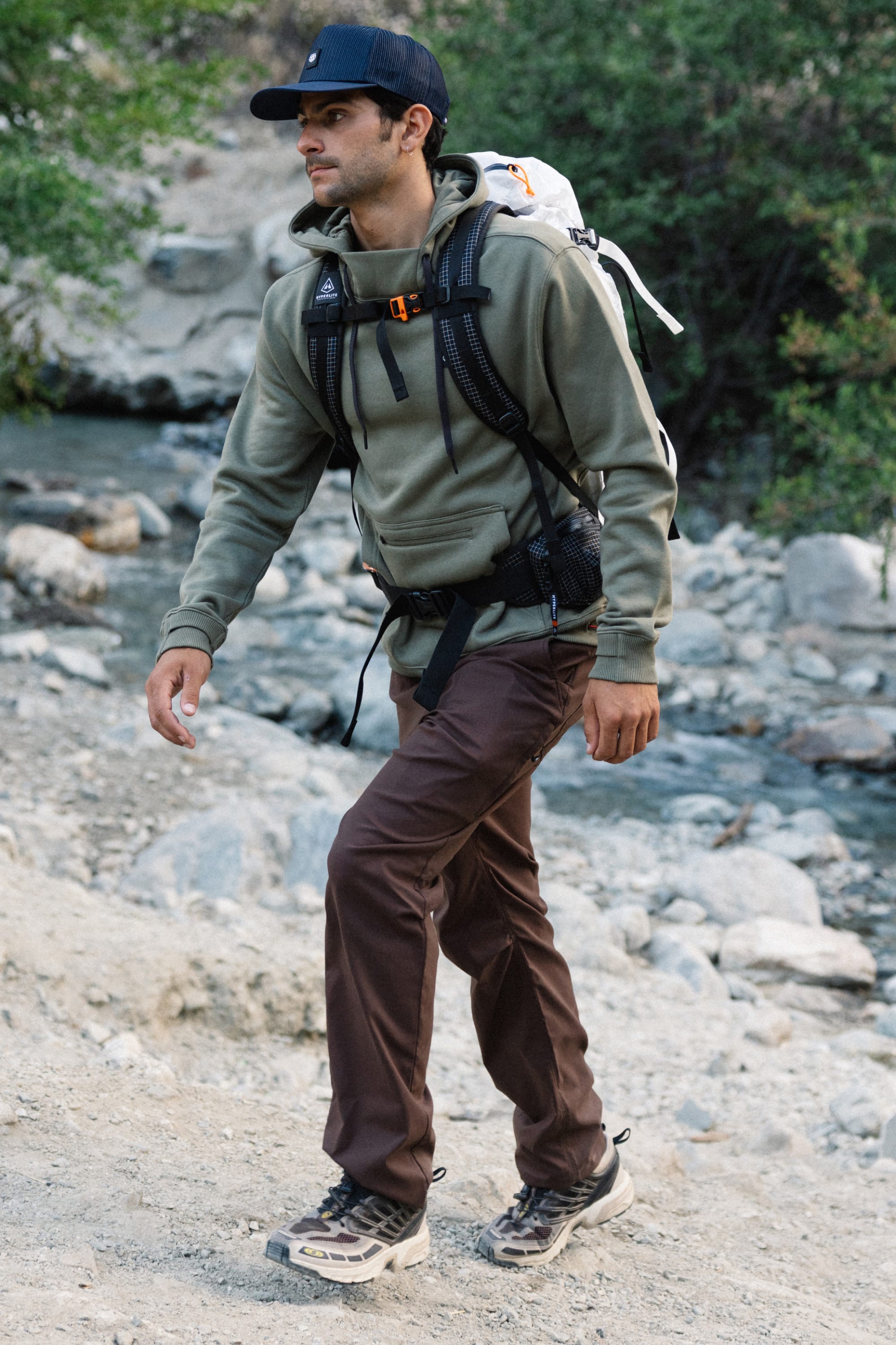 A man wearing a green hoodie, 686 Men's Everywhere® Pant - Relaxed Fit by 686, hiking boots, and a backpack walks on a rocky trail near a creek, surrounded by greenery.