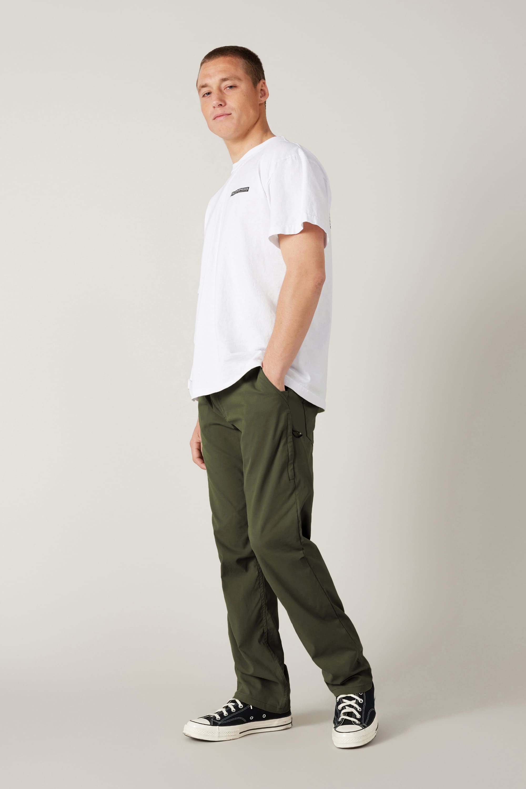 A person stands in a relaxed pose against a plain light background, wearing a breathable white T-shirt, black-and-white sneakers, and 686 Men's Everywhere® Pant - Relaxed Fit by 686 in olive green, with one hand in their pocket.