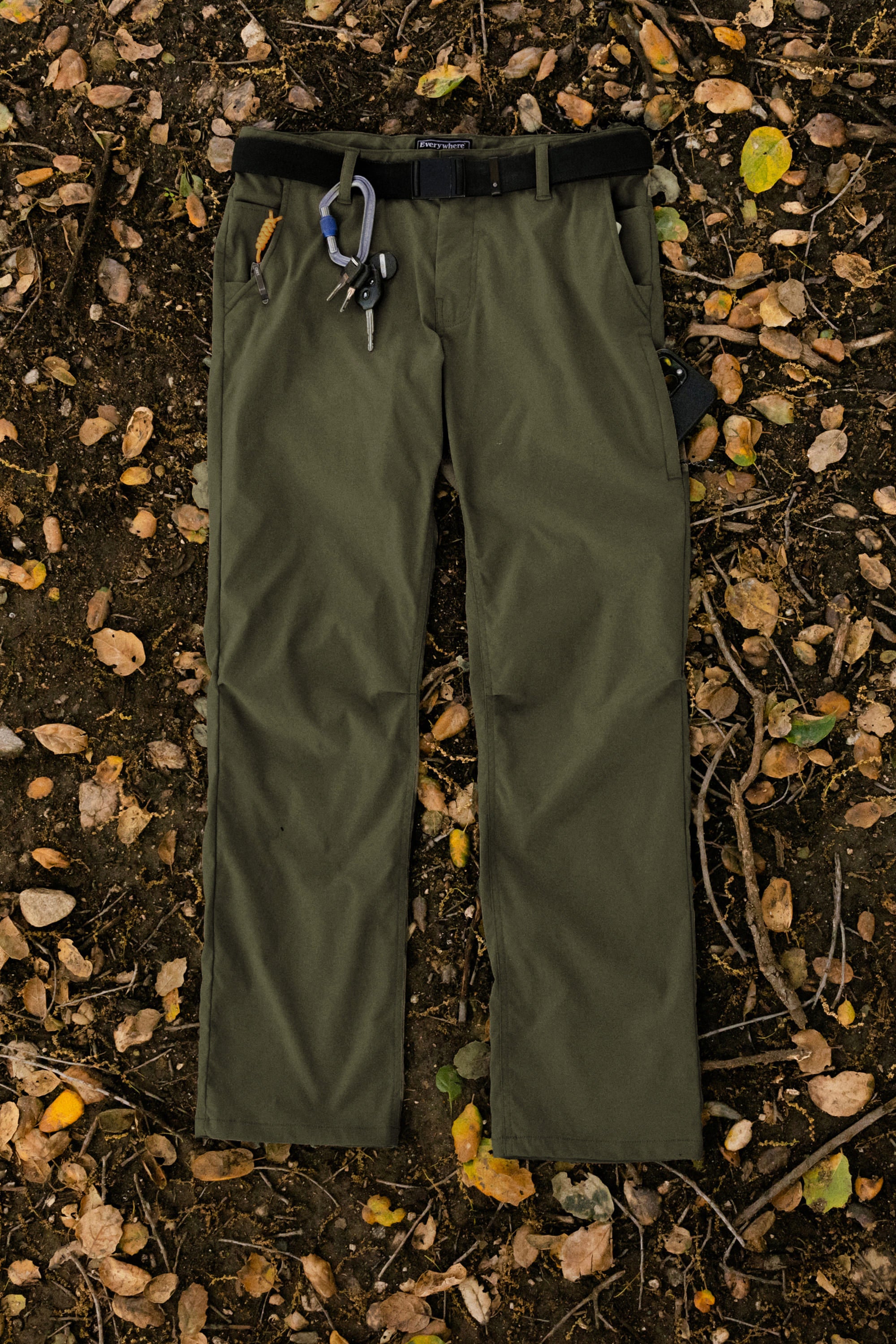 A pair of 686 Men's Everywhere® Pant - Relaxed Fit in olive green, featuring a black belt, keys, and a carabiner on the waistband, is laid flat on leaf-littered ground.
