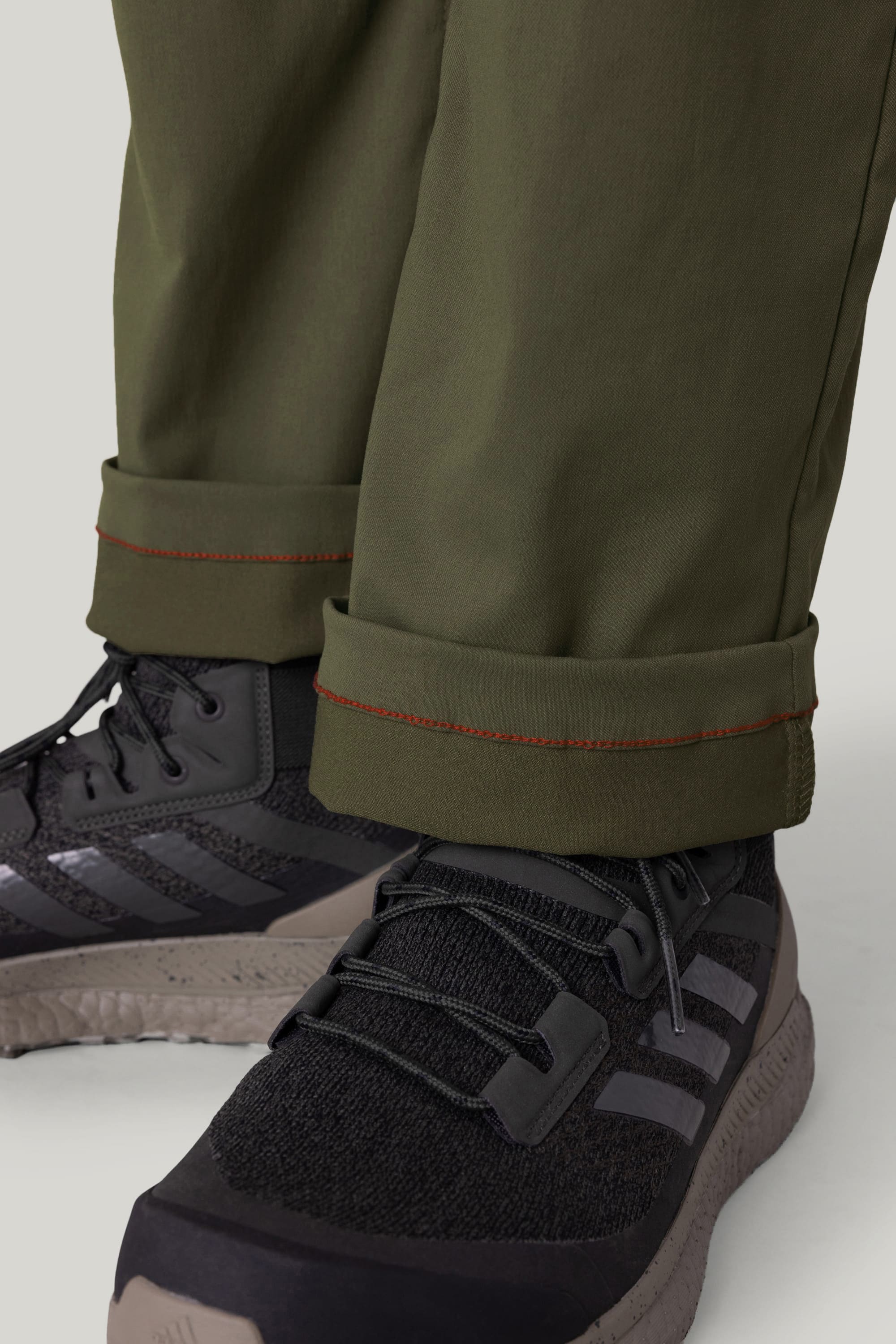Close-up of a person wearing 686 Men's Everywhere® Pant - Relaxed Fit in olive green, featuring red stitching near the cuffs. Paired with black athletic shoes with gray accents and textured soles. Pants are made from breathable fabric.