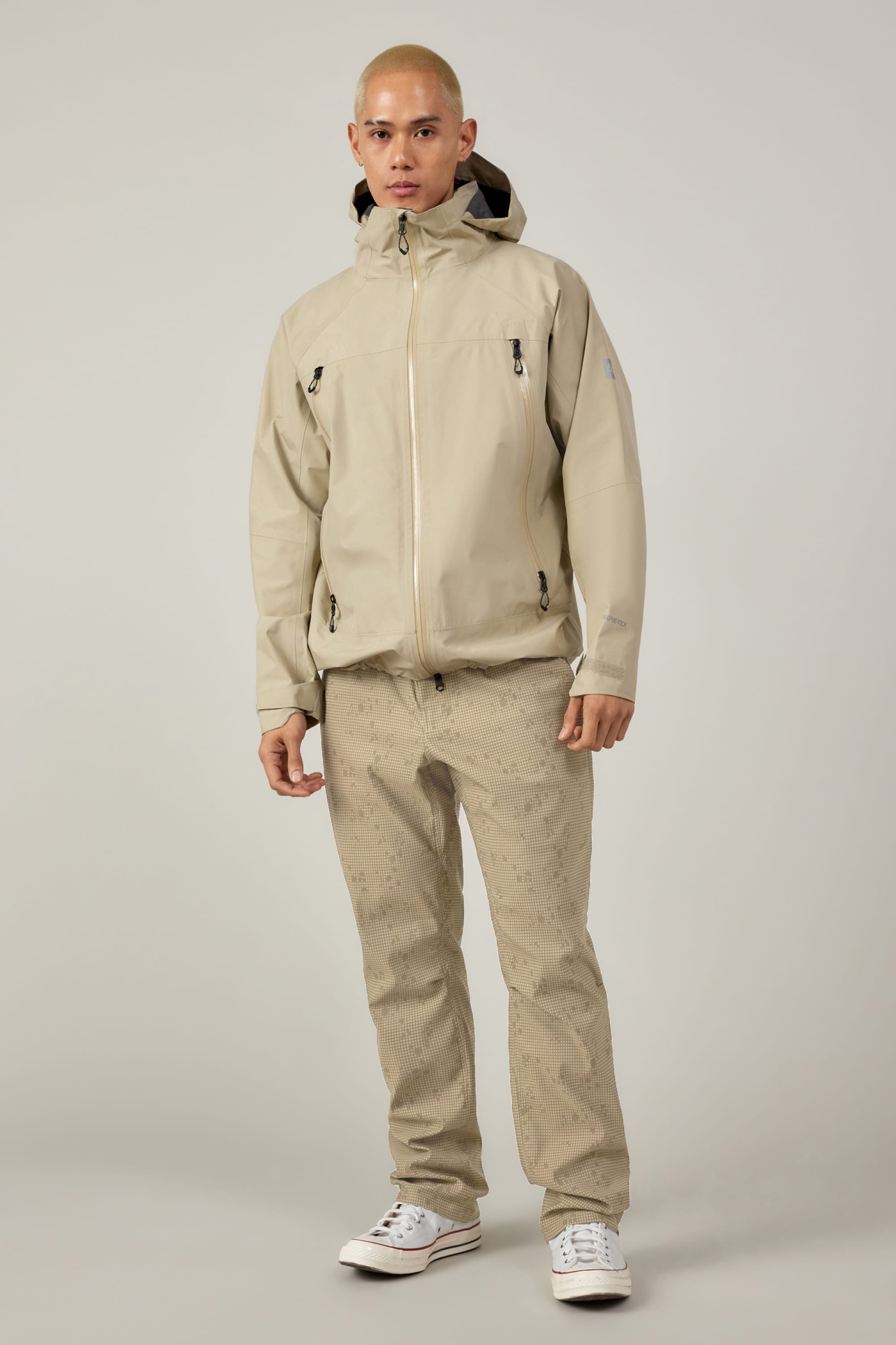 A person with short hair wears a beige hooded jacket, 686 Men's Everywhere® Pant - Relaxed Fit by 686, and white sneakers while standing against a plain light background.
