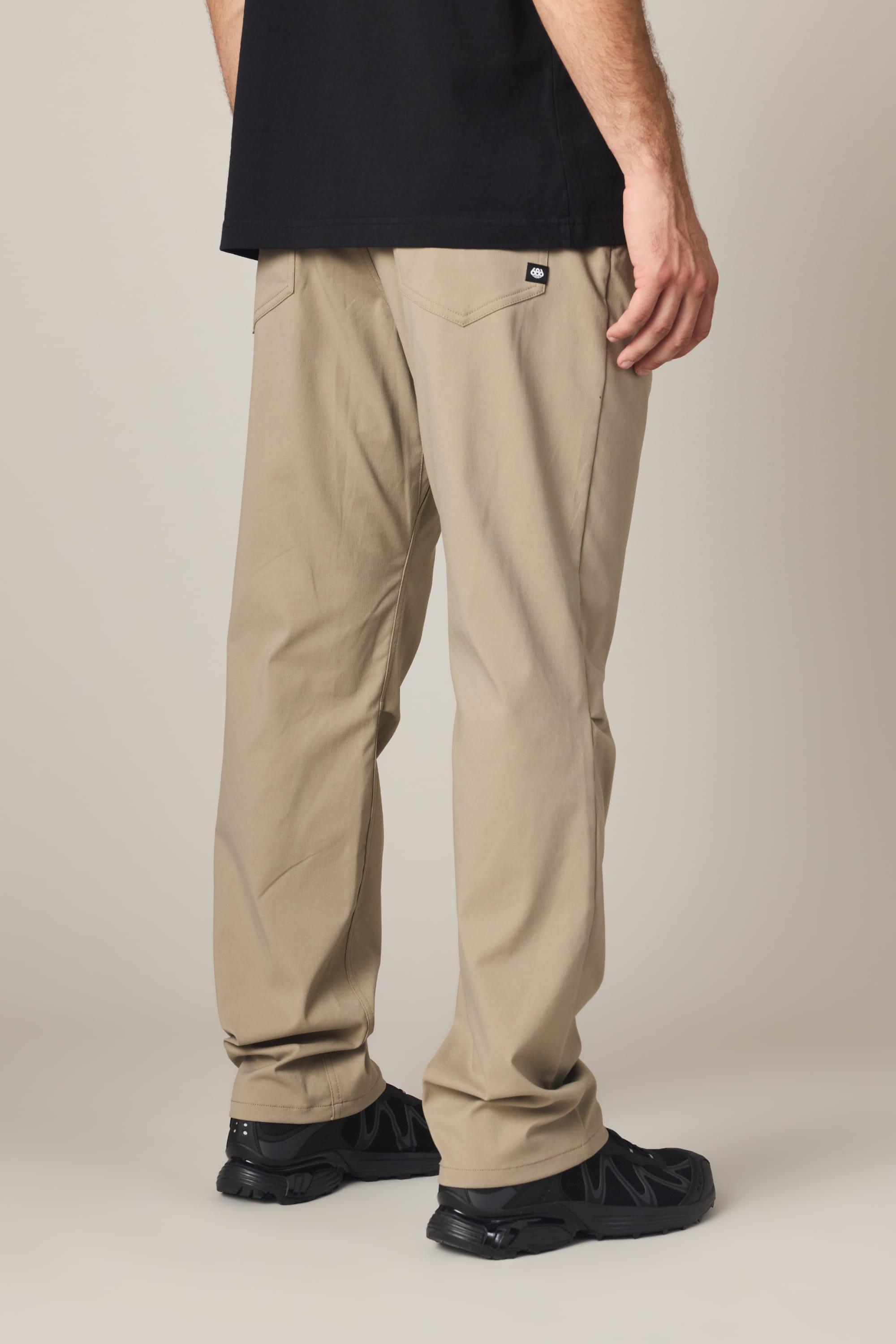 A person wearing the 686 Men's Everywhere® Pant - Relaxed Fit in beige, along with a black shirt and black shoes, stands facing away to display the back of the pants and legs against a plain background.