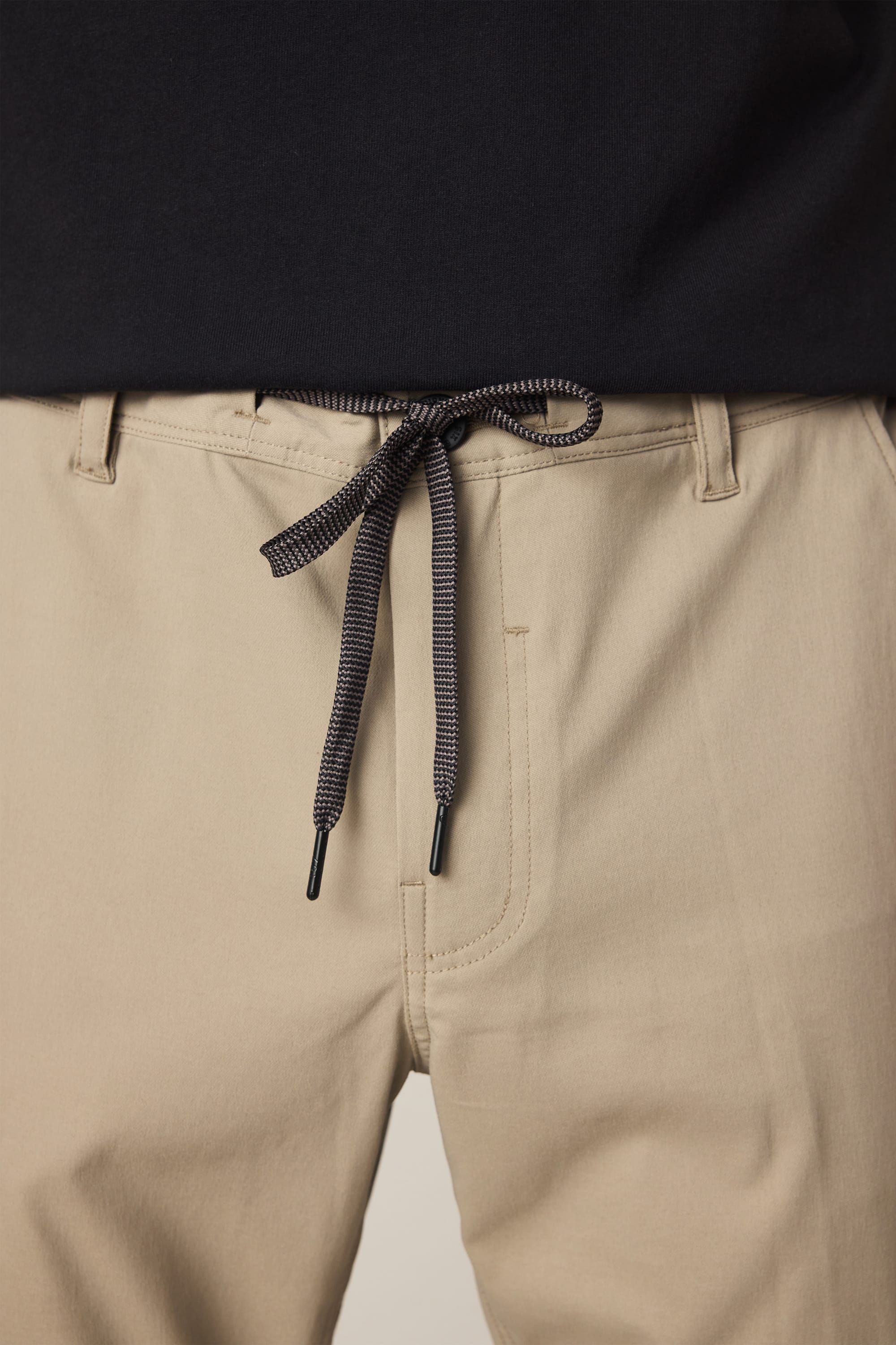 Close-up of a person wearing the 686 Men's Everywhere® Pant - Relaxed Fit in beige, featuring a black and white drawstring tie at the waistband, paired with a black top.