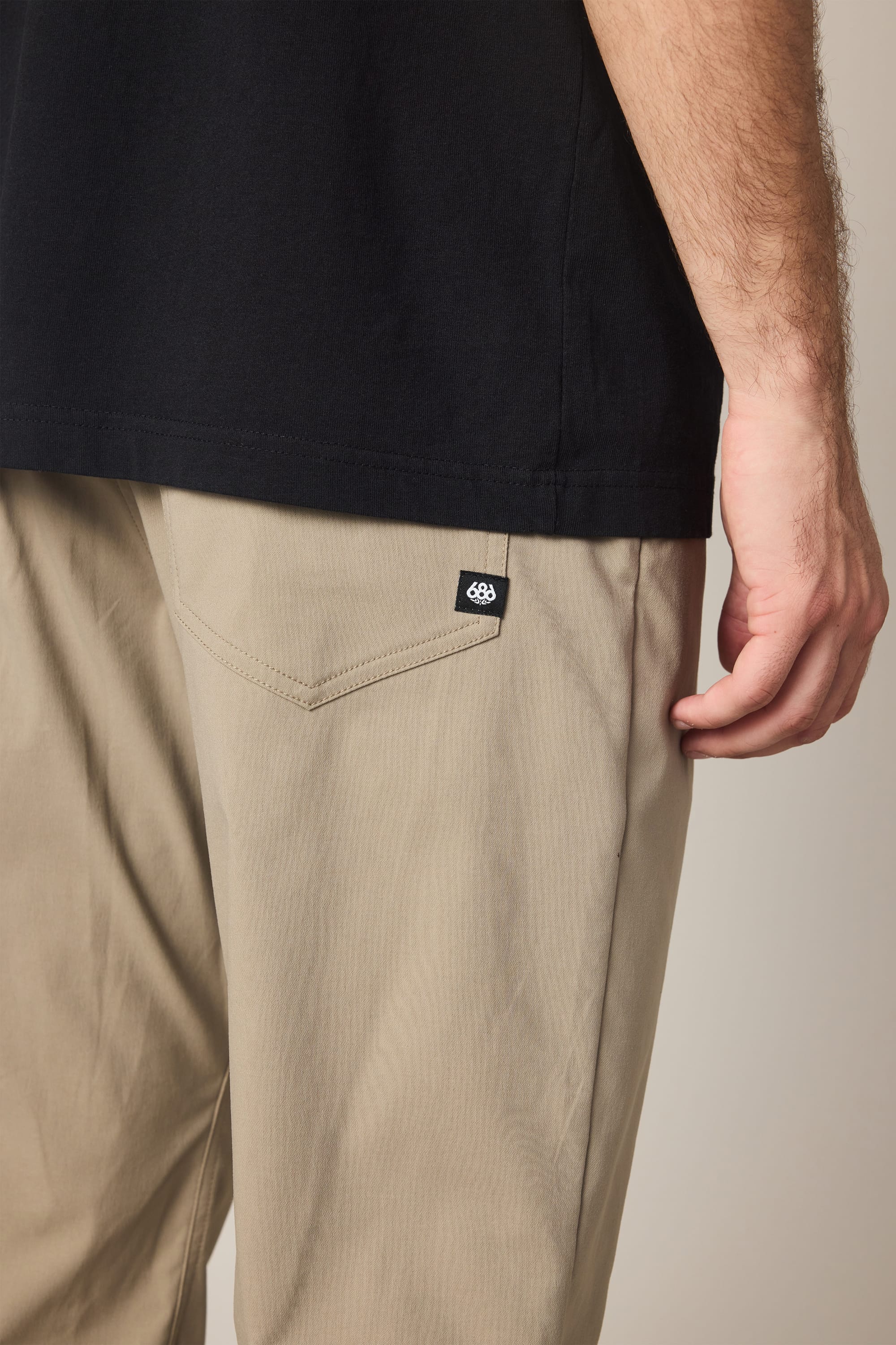 A person is wearing 686 Men's Everywhere® Pant - Relaxed Fit in beige, featuring a black tag near the back pocket, paired with a black shirt. Only the lower torso and right arm are visible.