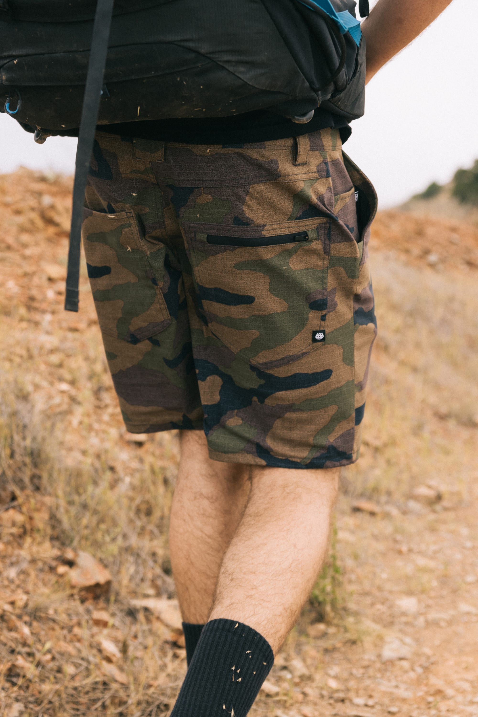 A person in 686 Men's Everywhere® Hybrid Short, black socks, and a backpack walks on a rocky dirt trail outdoors, with only their lower body visible.