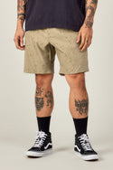 A person in tan checkered 686 Men's Everywhere® Hybrid Short, a black shirt, black socks, and black Vans sneakers, with tattoos on arms and legs, stands against a light background. The 686 shorts are breathable and stain-resistant for all-day comfort.