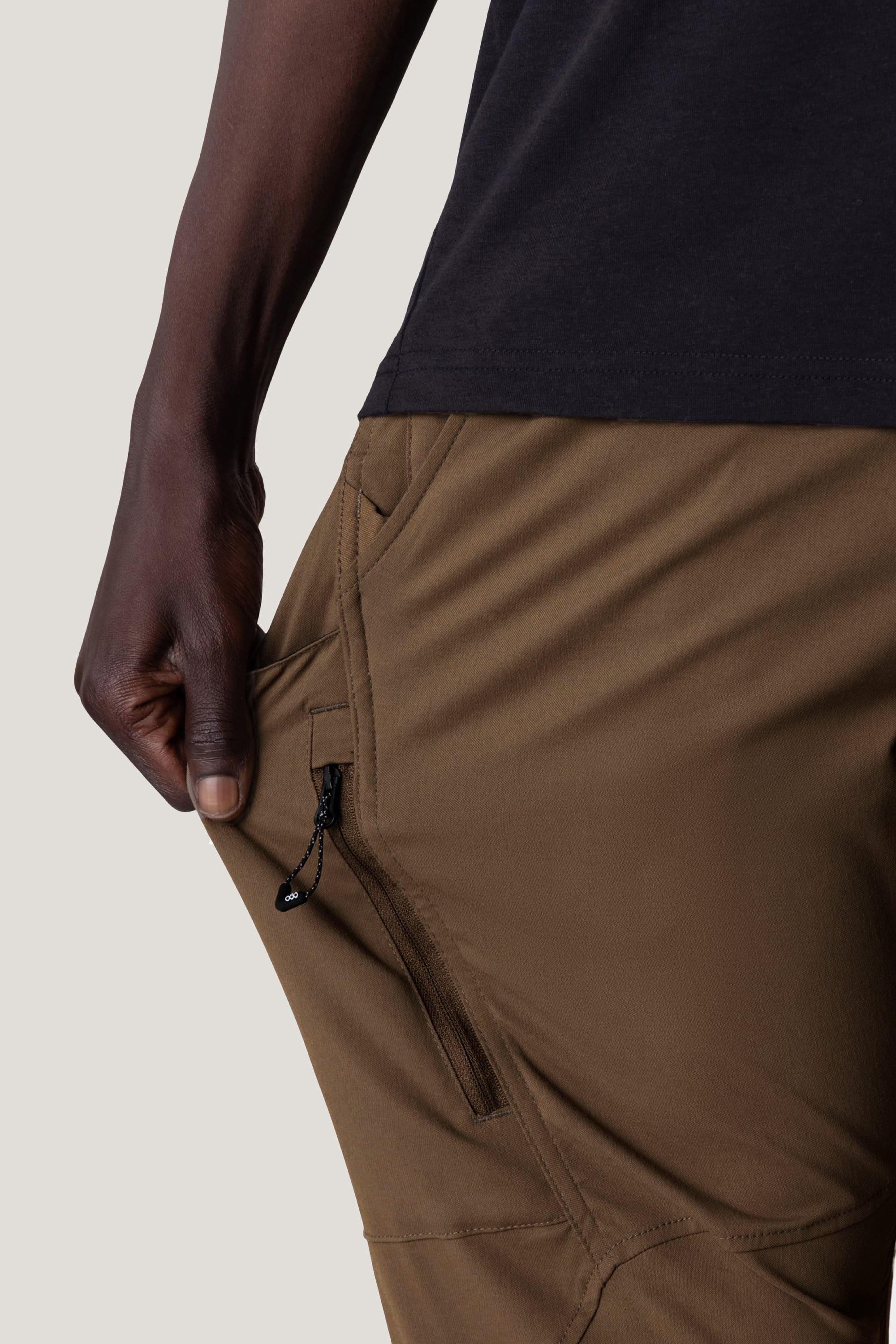 A person in 686 Men's Anything Cargo Pant - Slim Fit (brown) and a black shirt pulls at a zippered pocket, highlighting the 13-pocket design and breathable fabric from 686.