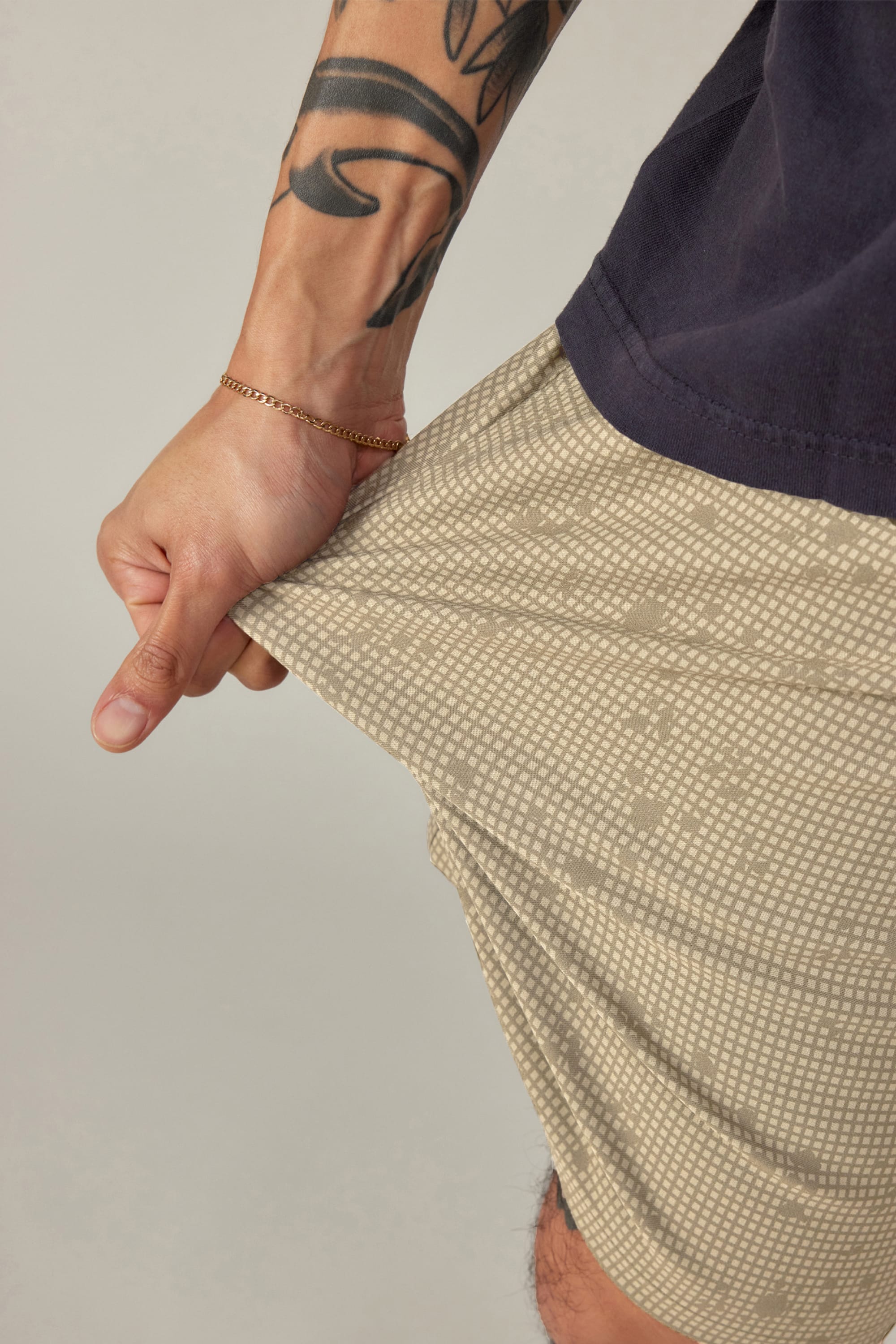 A tattooed arm with a gold bracelet stretches the fabric of 686 Men's Everywhere® Hybrid Short - Relaxed Fit while wearing a dark shirt, highlighting these beige patterned, breathable shorts from 686.