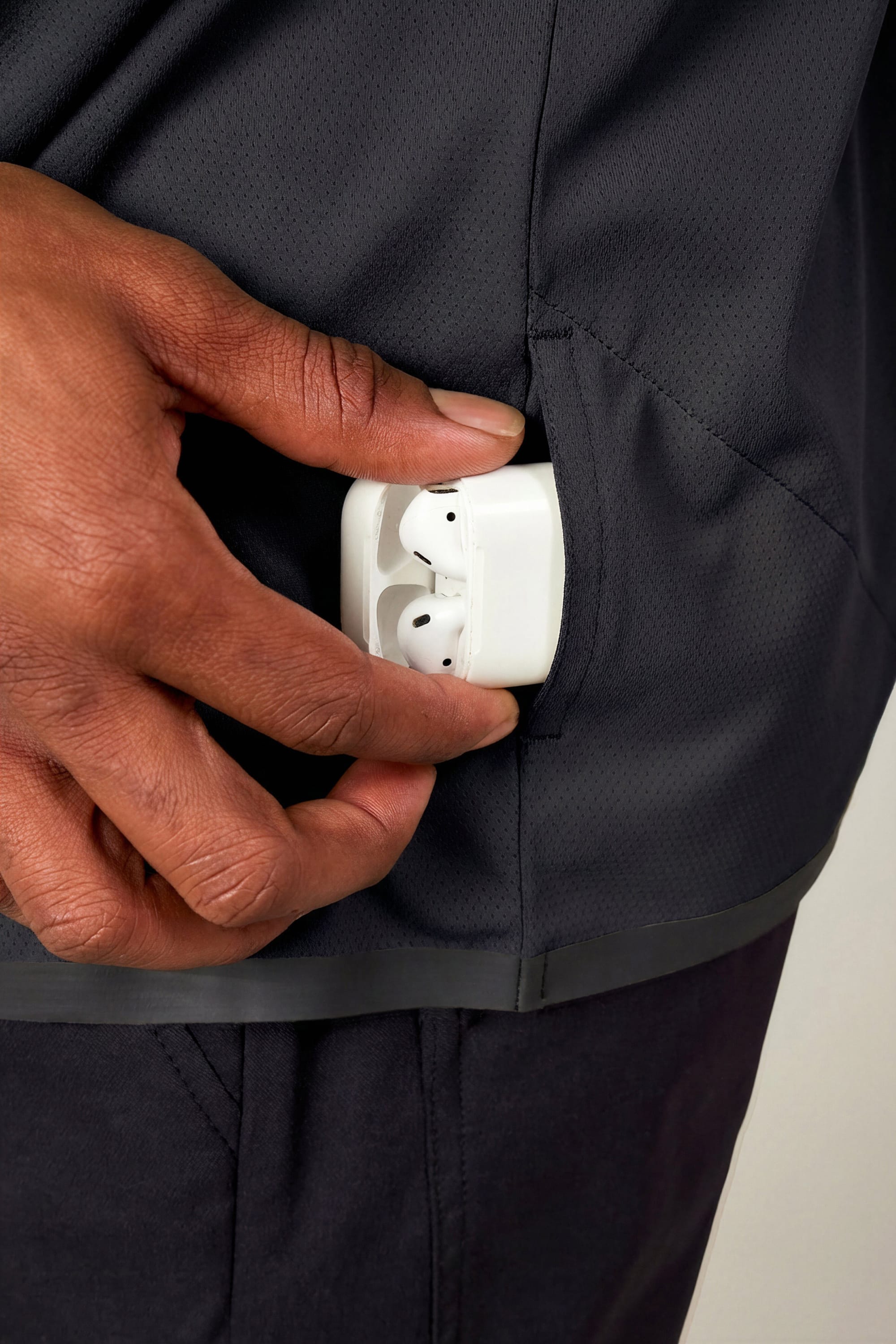 A person places a white wireless earbud case into the side pocket of the 686 Men's Let's Go Tech Hoody, a lightweight black jacket ideal for sun protection.