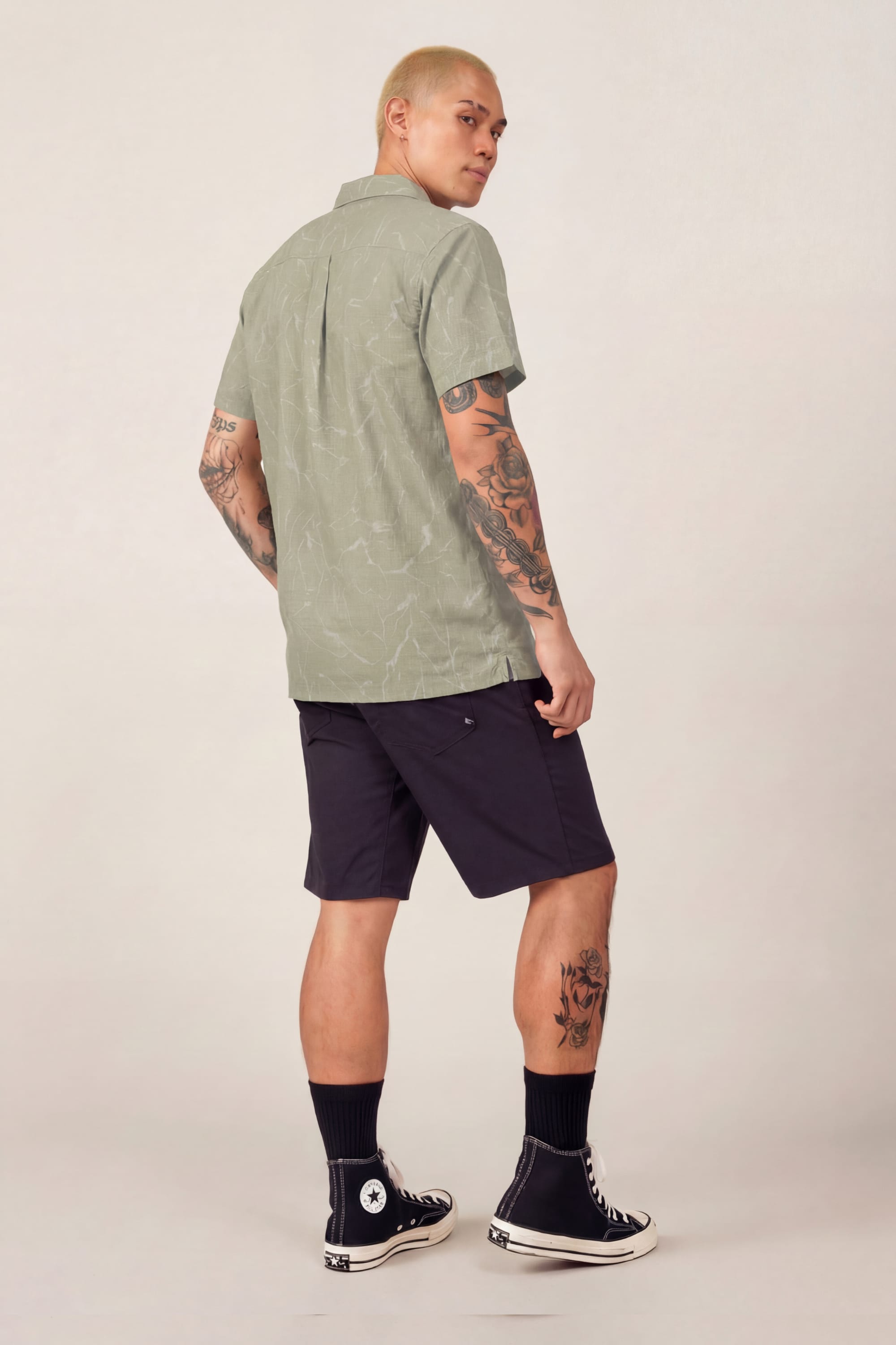 A person with short blond hair and visible tattoos wears the 686 Men's Nomad Perforated Button Down Shirt in light green, dark shorts, black socks, and black Converse sneakers, standing and looking over their shoulder.