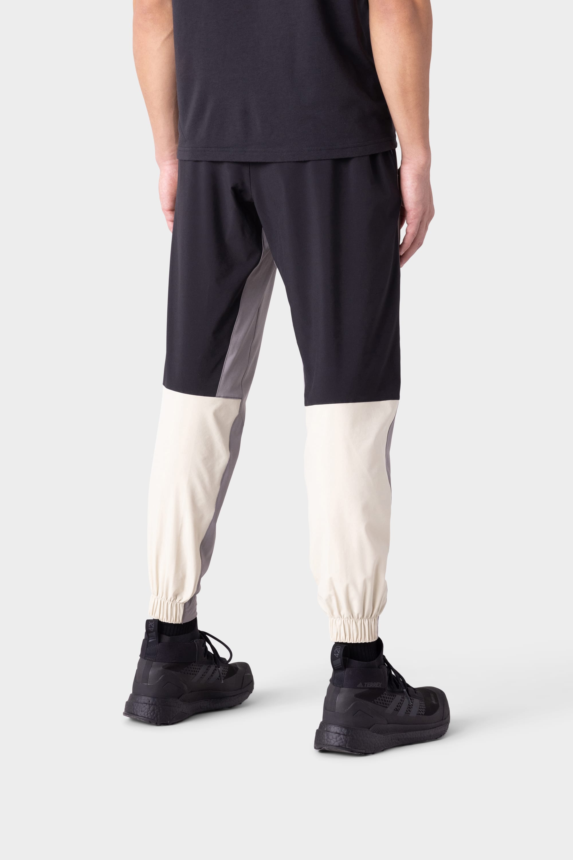 A person stands facing away, wearing a black shirt and 686 Men's ATP Stretch Performance Pant by 686, which features elastic cuffs, black upper sections, gray side panels, and off-white lower legs. Black sneakers complete the look.