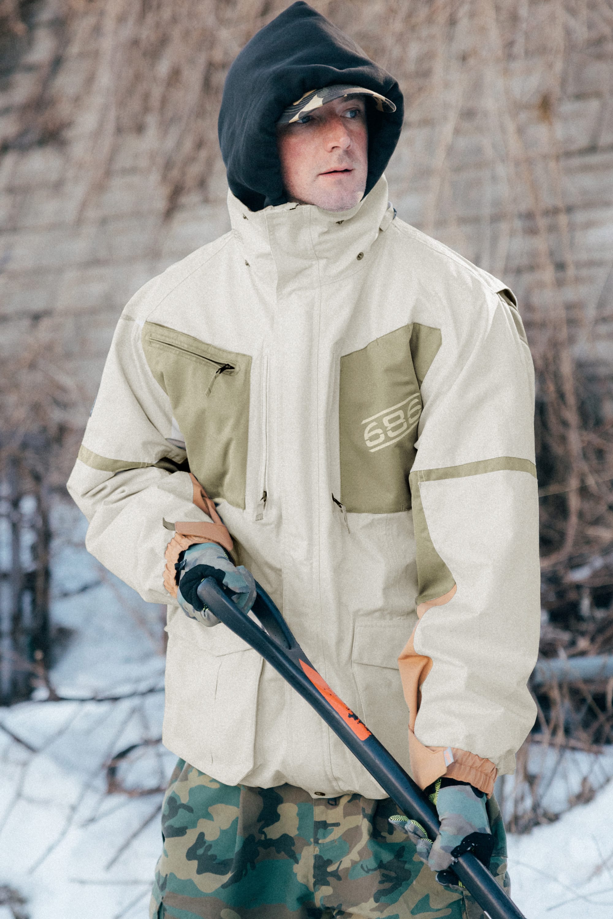 Wearing a 686 Mens 2001 Transfer Jacket and camouflage pants, a person holds a ski pole while standing outdoors in the snow, with bare branches and a stone wall in the background.