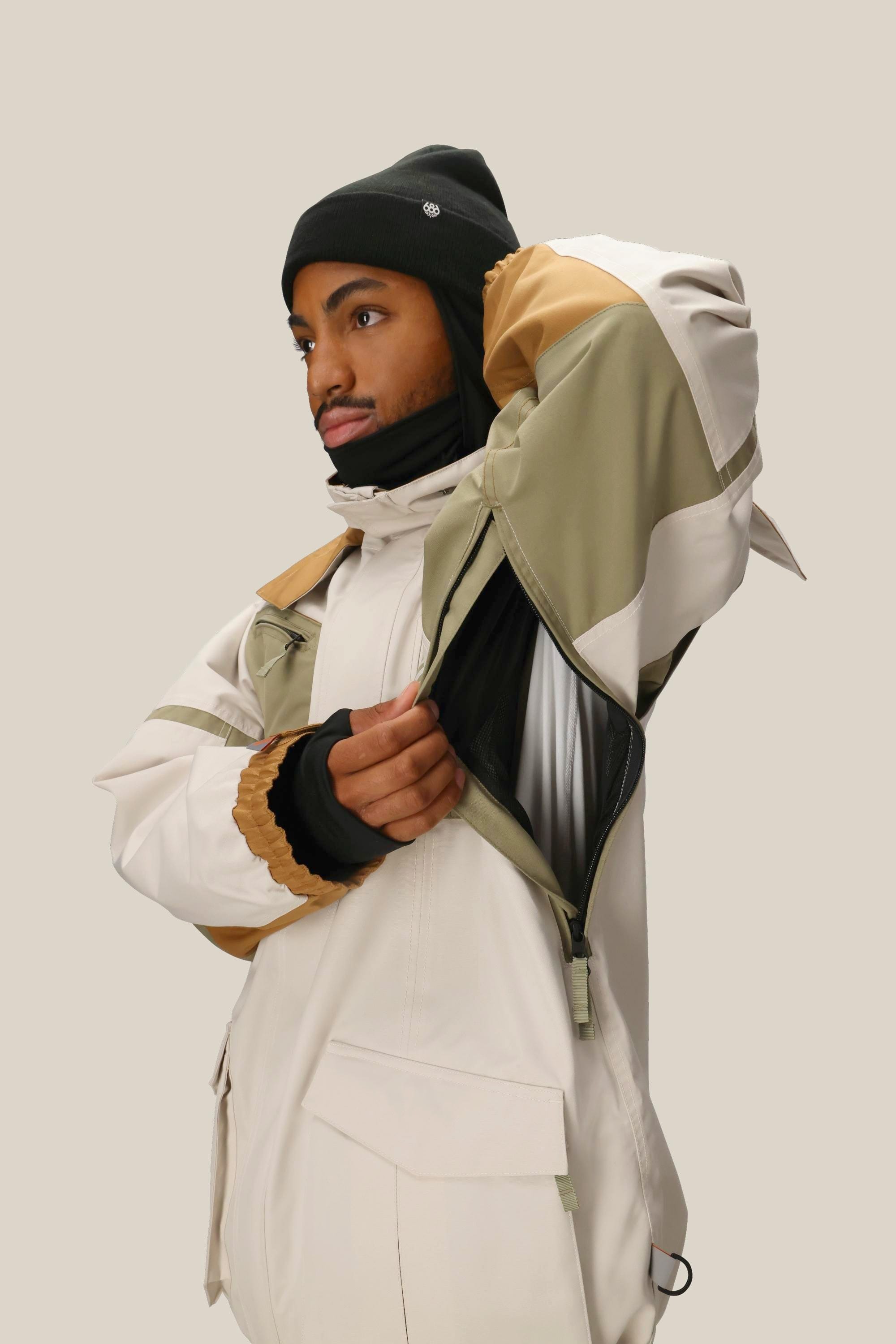 A person in the 686 Mens 2001 Transfer Jacket in beige and olive green, paired with a black beanie and gloves, unzips a side vent while standing against a plain light background.
