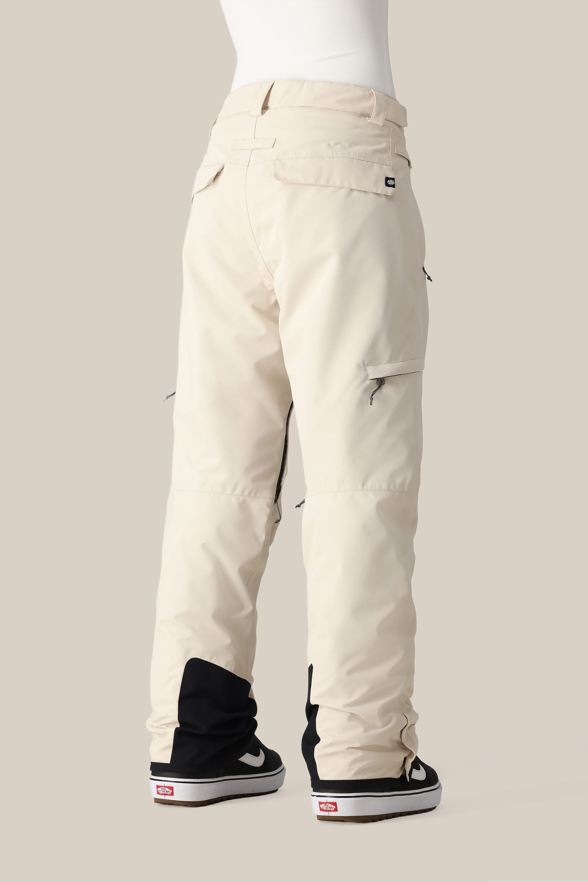 A person wears 686 Women's SMARTY® 3-in-1 Cargo Pants in cream with side pockets, black ankle reinforcements, and a removable sweatpant liner. The 686 pants offer three ways to wear and are styled with black and white skate shoes against a beige background.