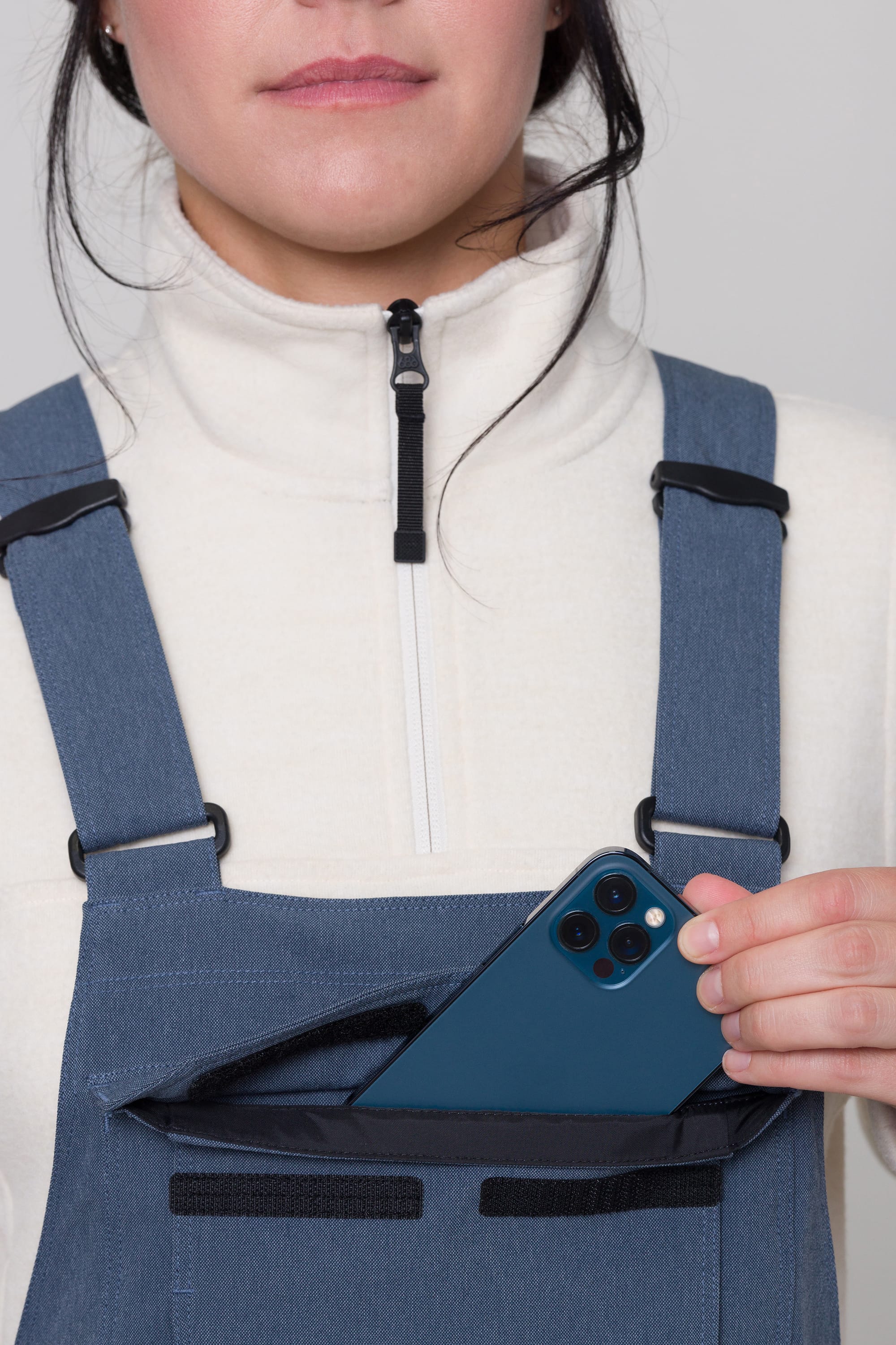 A person wearing the 686 Womens Black Magic Insulated Bib by 686, known for extra pocketing, slips a dark blue smartphone into the front pocket. Only their lower face and hand are visible.