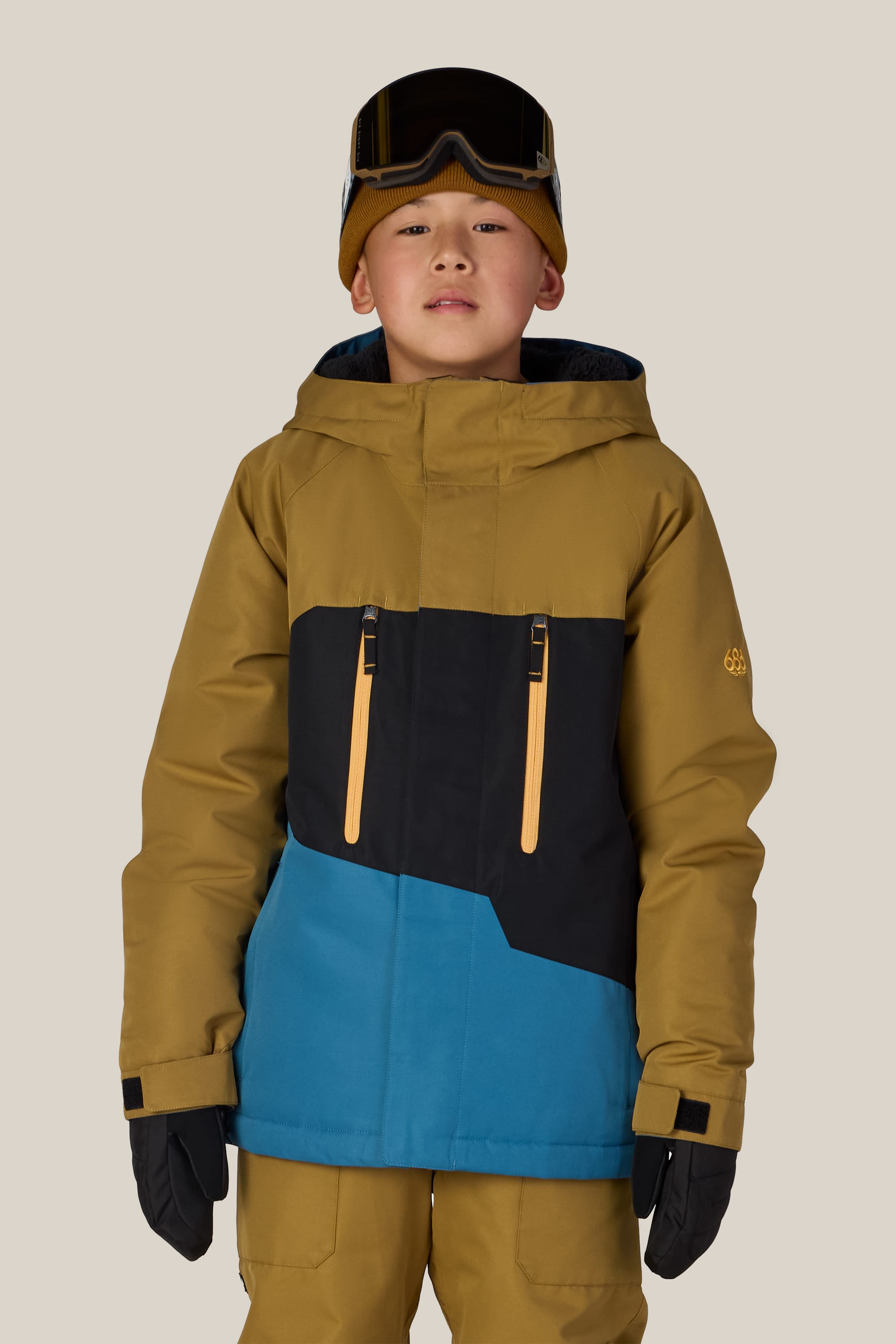A child wearing the 686 Boys’ Geo™ Insulated Jacket by 686 in mustard yellow and blue, along with matching pants, black gloves, a brown beanie, and ski goggles on their head stands facing forward against a plain background.
