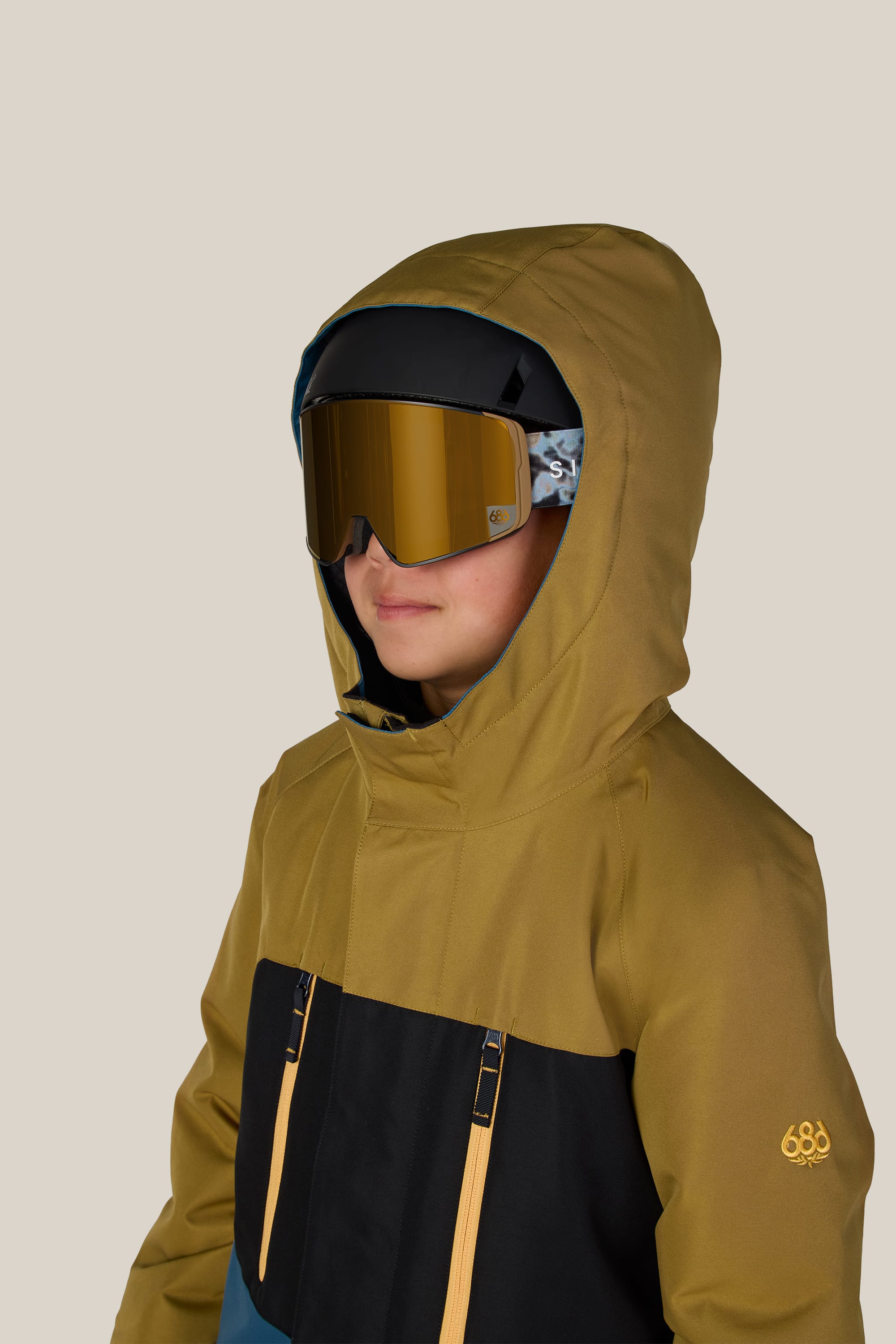 A child wearing the 686 Boys’ Geo™ Insulated Jacket in mustard yellow and black by 686, along with reflective ski goggles and a helmet, standing against a plain light background.
