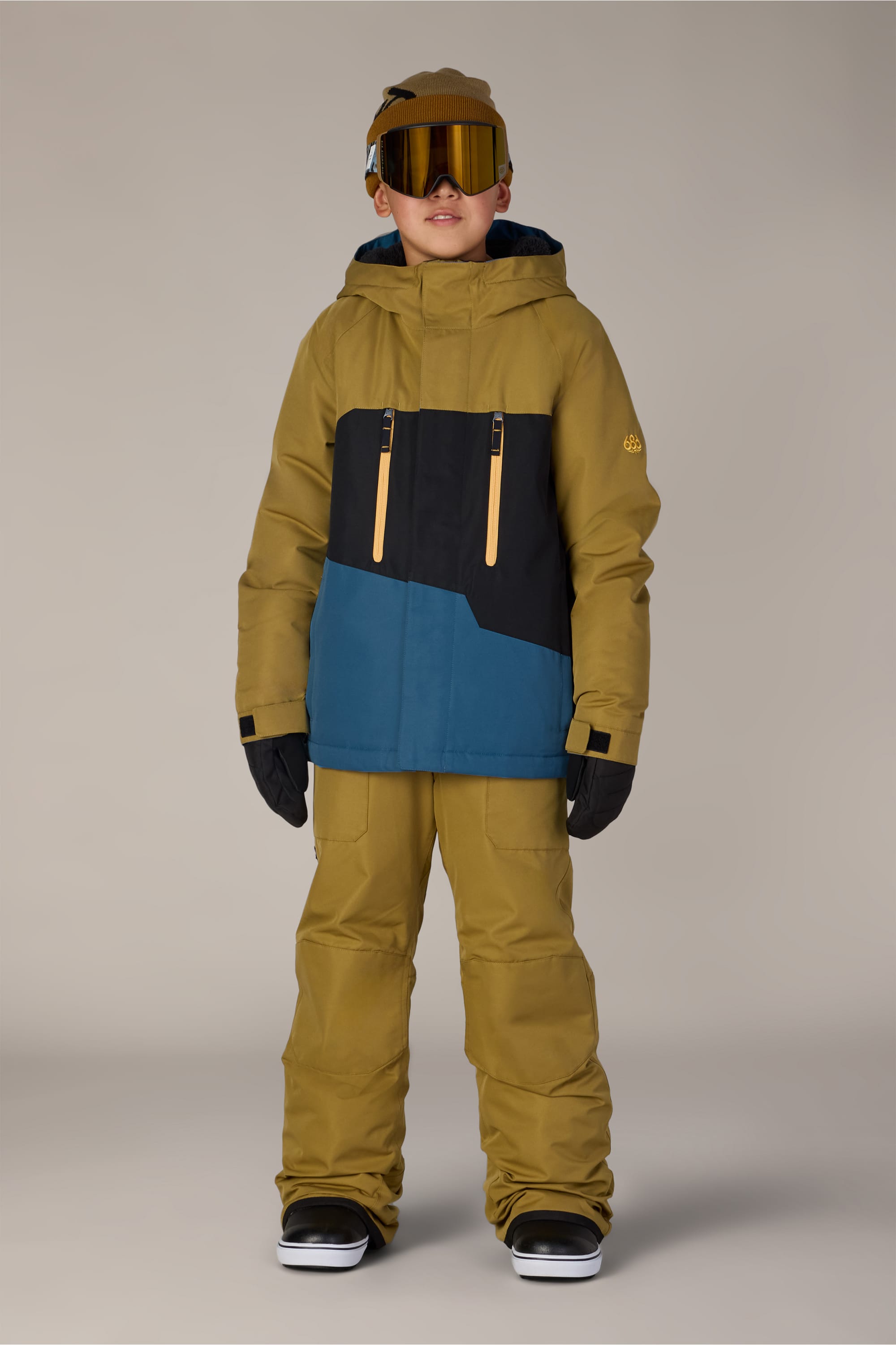 A child stands facing forward against a plain light gray background, wearing the 686 Boys’ Geo™ Insulated Jacket in mustard yellow and blue from 686, paired with matching pants, black gloves, black boots, brown ski goggles, and a brown cap.
