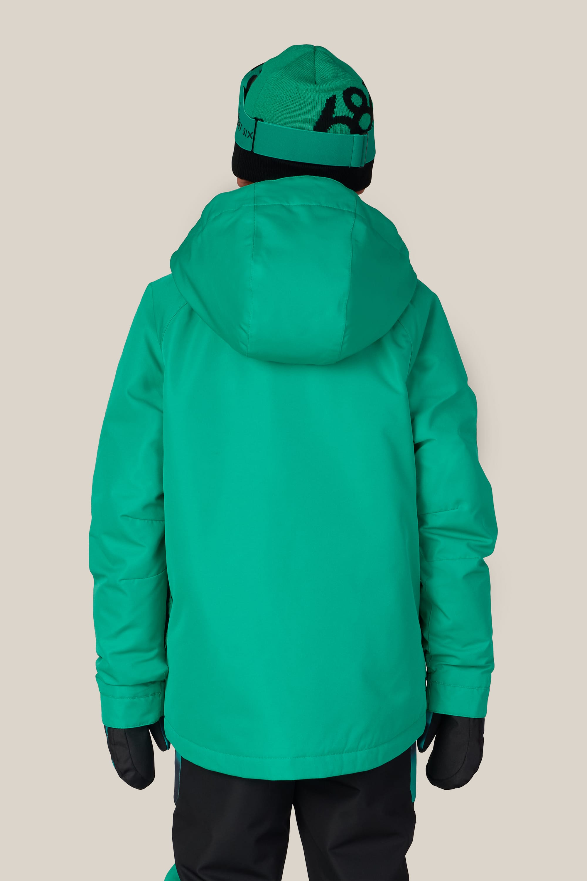 A child is shown from the back wearing the 686 Boys’ Geo™ Insulated Jacket in a bright teal color, along with a matching hat and black gloves, standing against a plain beige background.
