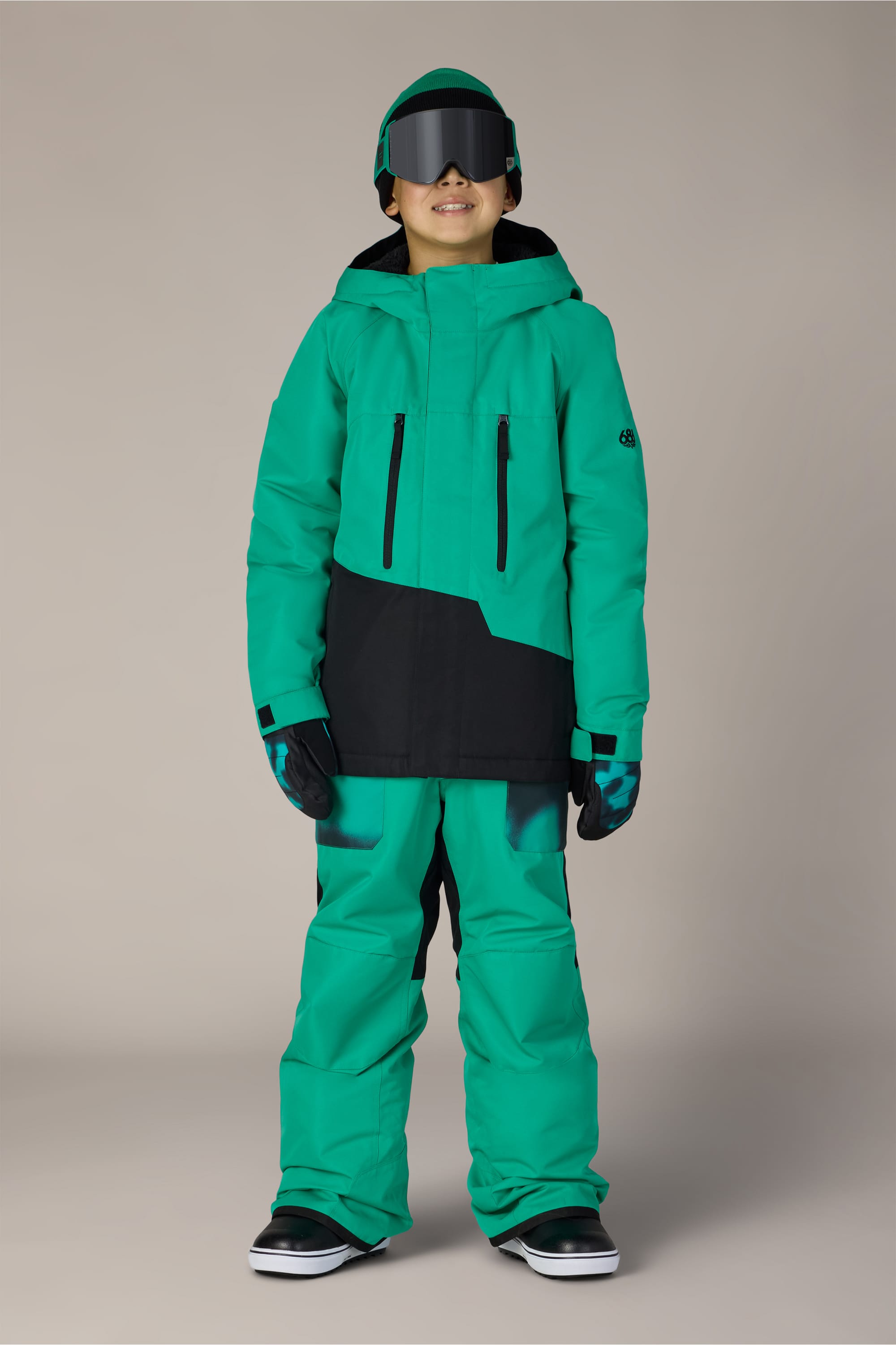 A child wearing the 686 Boys’ Geo™ Insulated Jacket by 686, along with matching pants, gloves, helmet, and goggles, stands facing forward and smiling against a plain background.