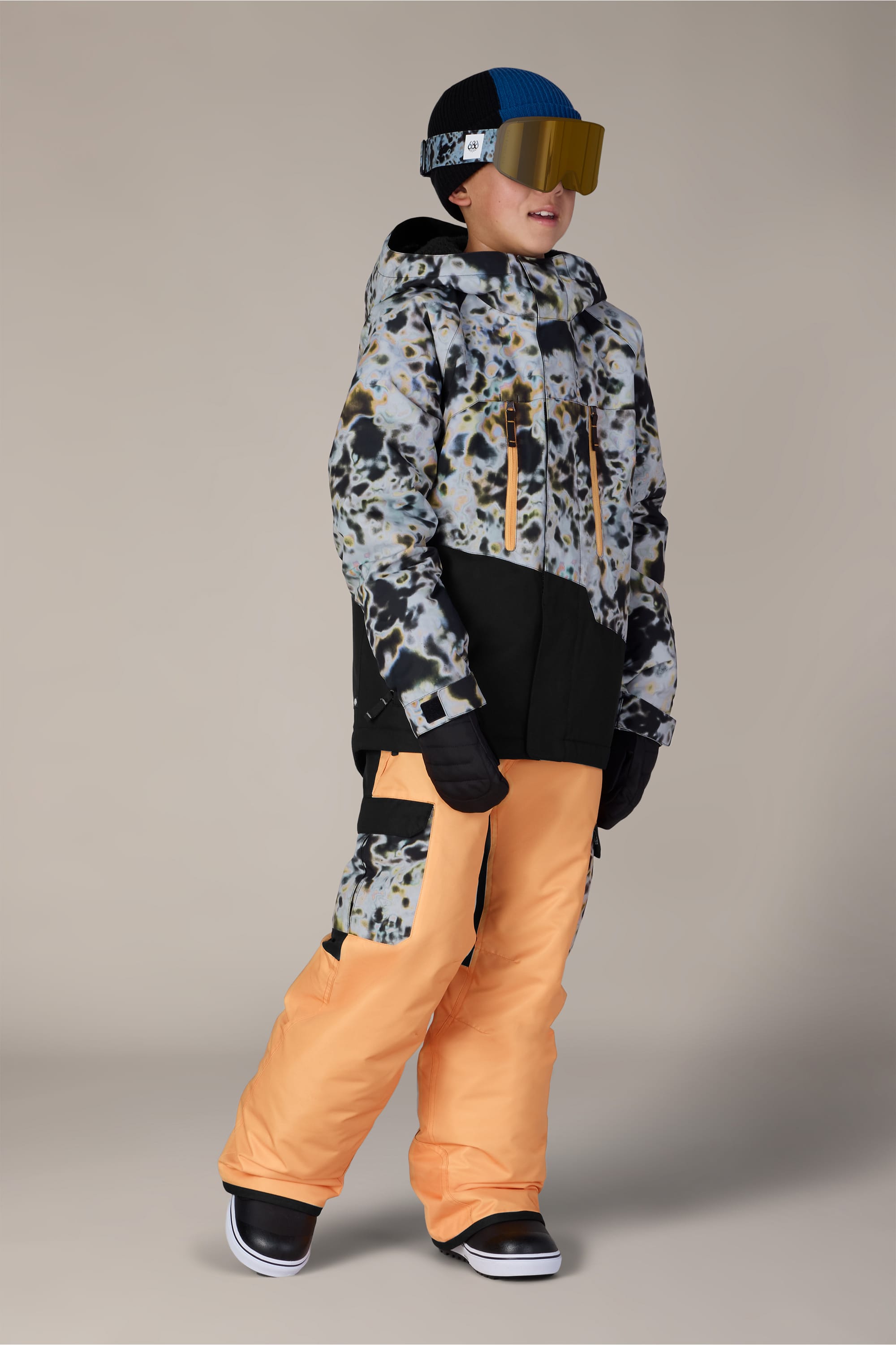 A child wearing the 686 Boys’ Geo™ Insulated Jacket by 686, paired with orange snow pants, black gloves, boots, a blue-and-black hat, and yellow ski goggles stands against a plain background.