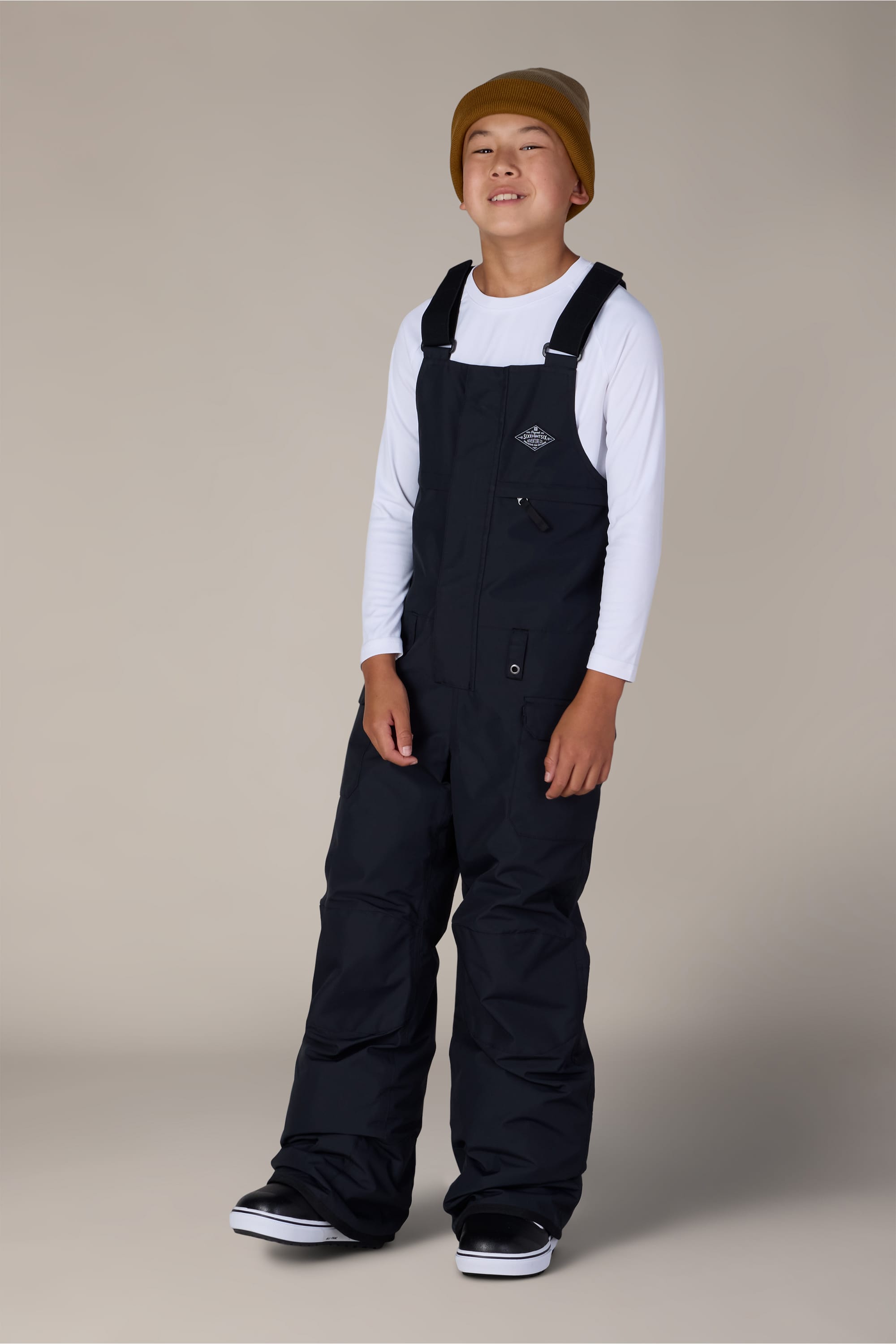 A smiling young person wears the 686 Boys’ Frontier™ Insulated Bib by 686, paired with a yellow beanie and a white long-sleeve shirt, standing against a plain light beige background.
