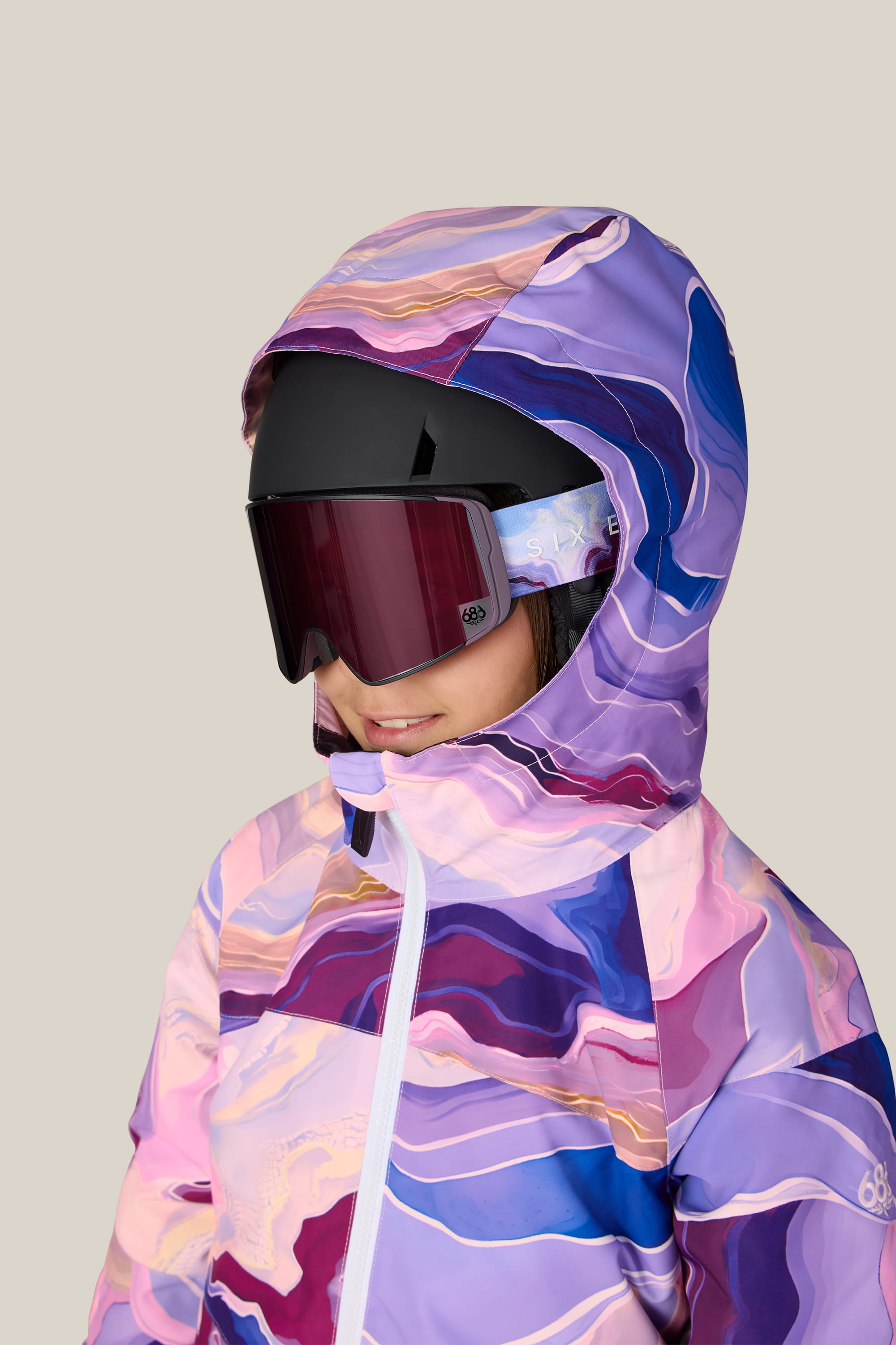 A person wearing the 686 Girls’ Athena™ Insulated Jacket—a purple and pink patterned ski jacket with a hood—paired with a black ski helmet and red-tinted ski goggles, against a light beige background.