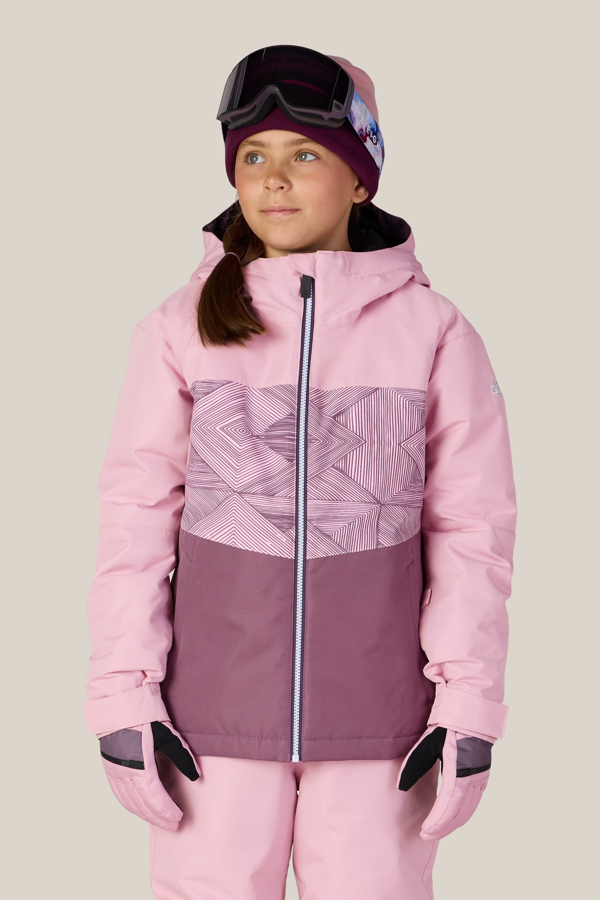 A young girl wearing the 686 Girls’ Athena™ Insulated Jacket by 686, along with matching gloves, snow pants, a beanie, and ski goggles on her forehead, stands against a light beige background.