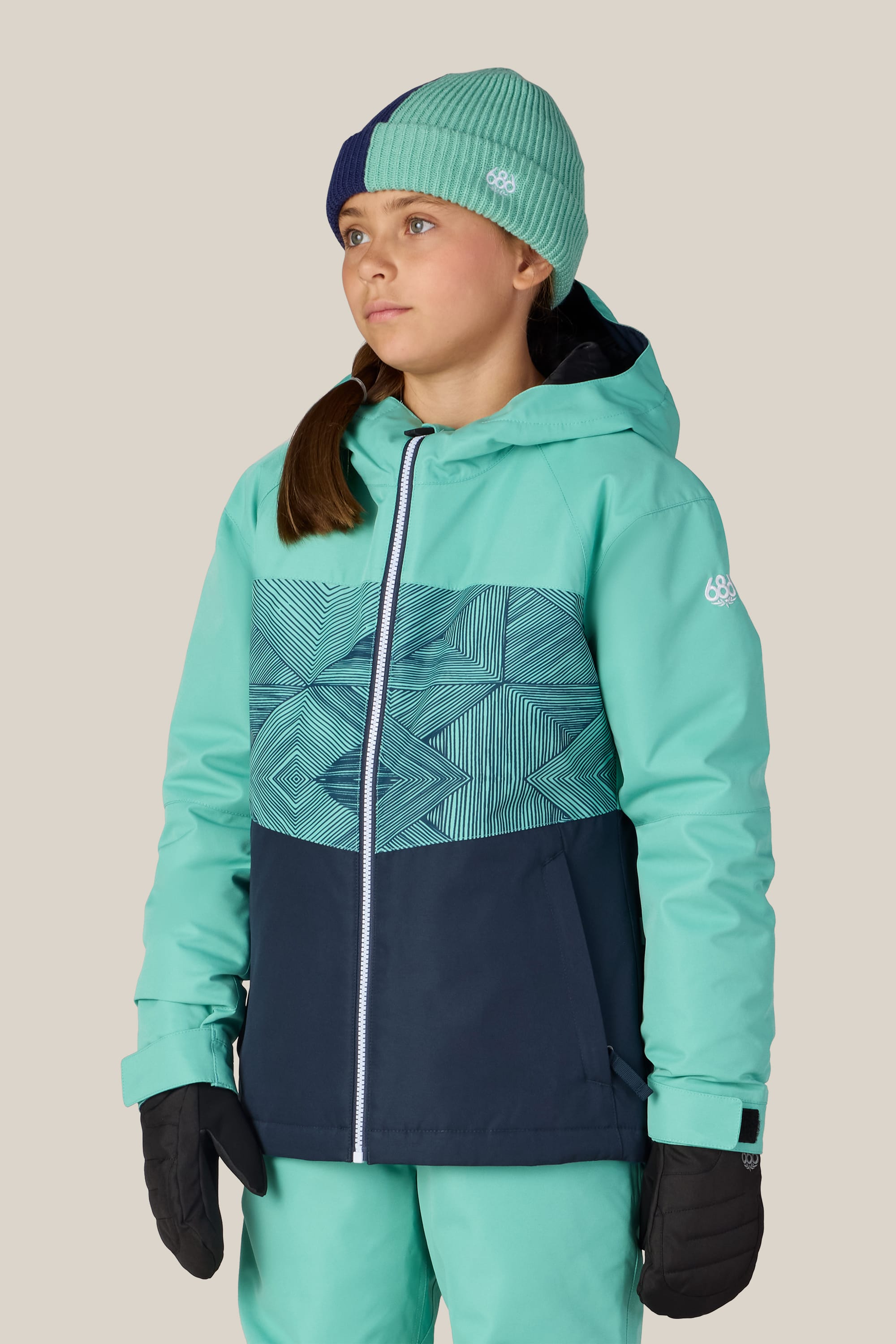 A girl wearing the 686 Girls’ Athena™ Insulated Jacket in teal and navy, paired with matching teal pants, black gloves, and a green beanie with a navy section, stands against a plain light background, looking slightly to the side.