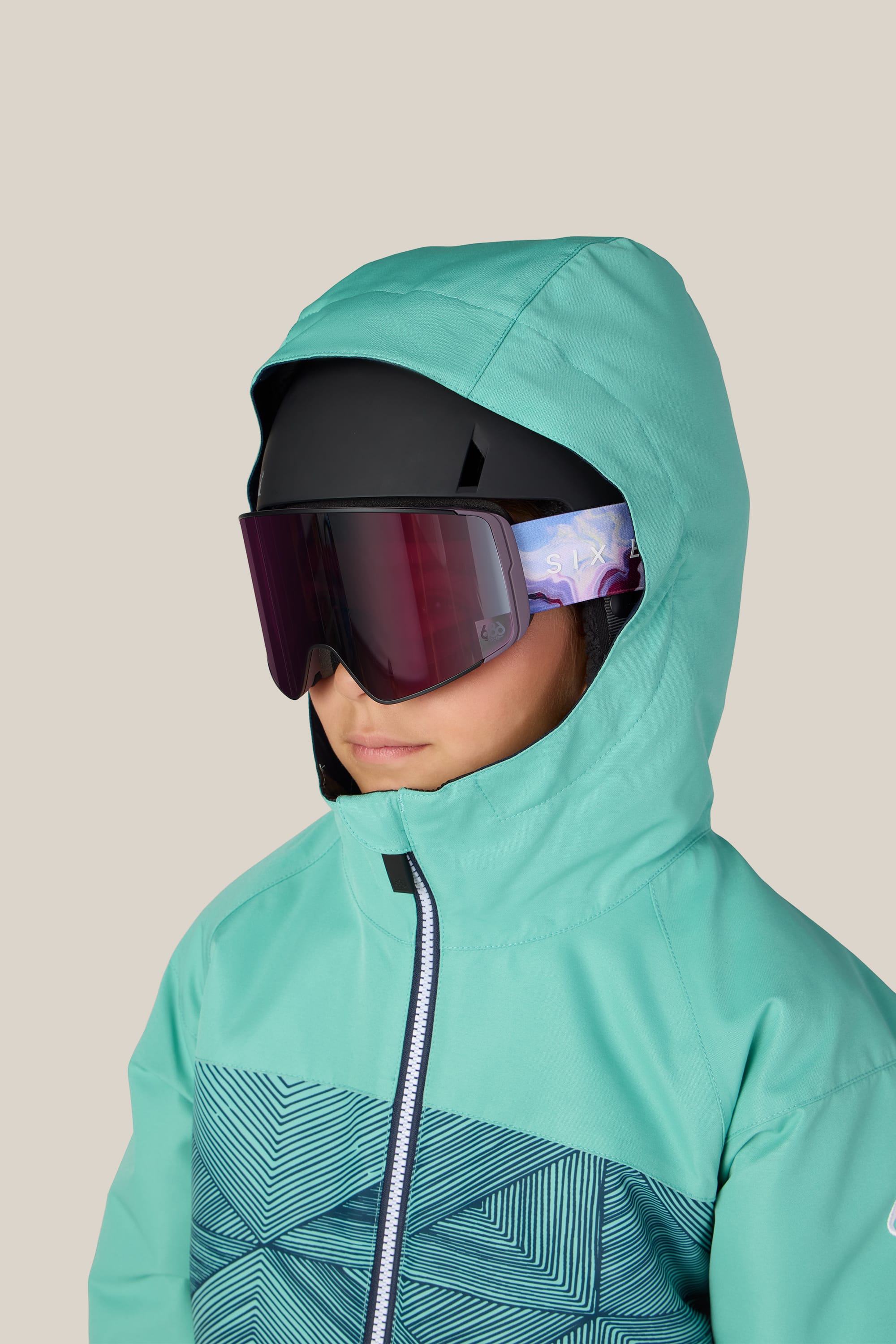 A child wearing the 686 Girls’ Athena™ Insulated Jacket in teal, paired with large dark ski goggles and a black helmet, faces slightly to the left against a plain beige background.