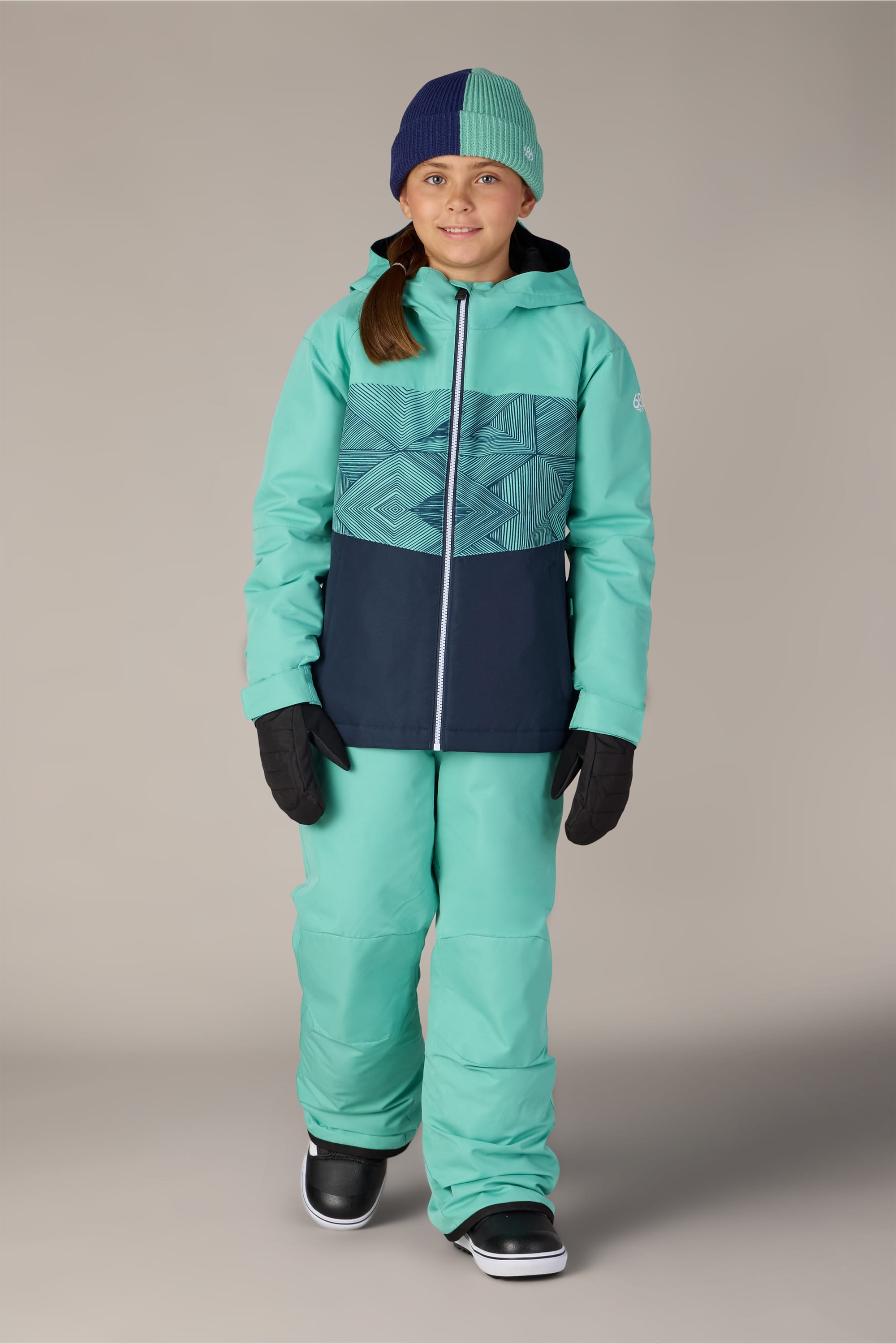 A child wearing the 686 Girls’ Athena™ Insulated Jacket by 686 stands facing forward, smiling against a plain background, paired with matching attire and accessories.