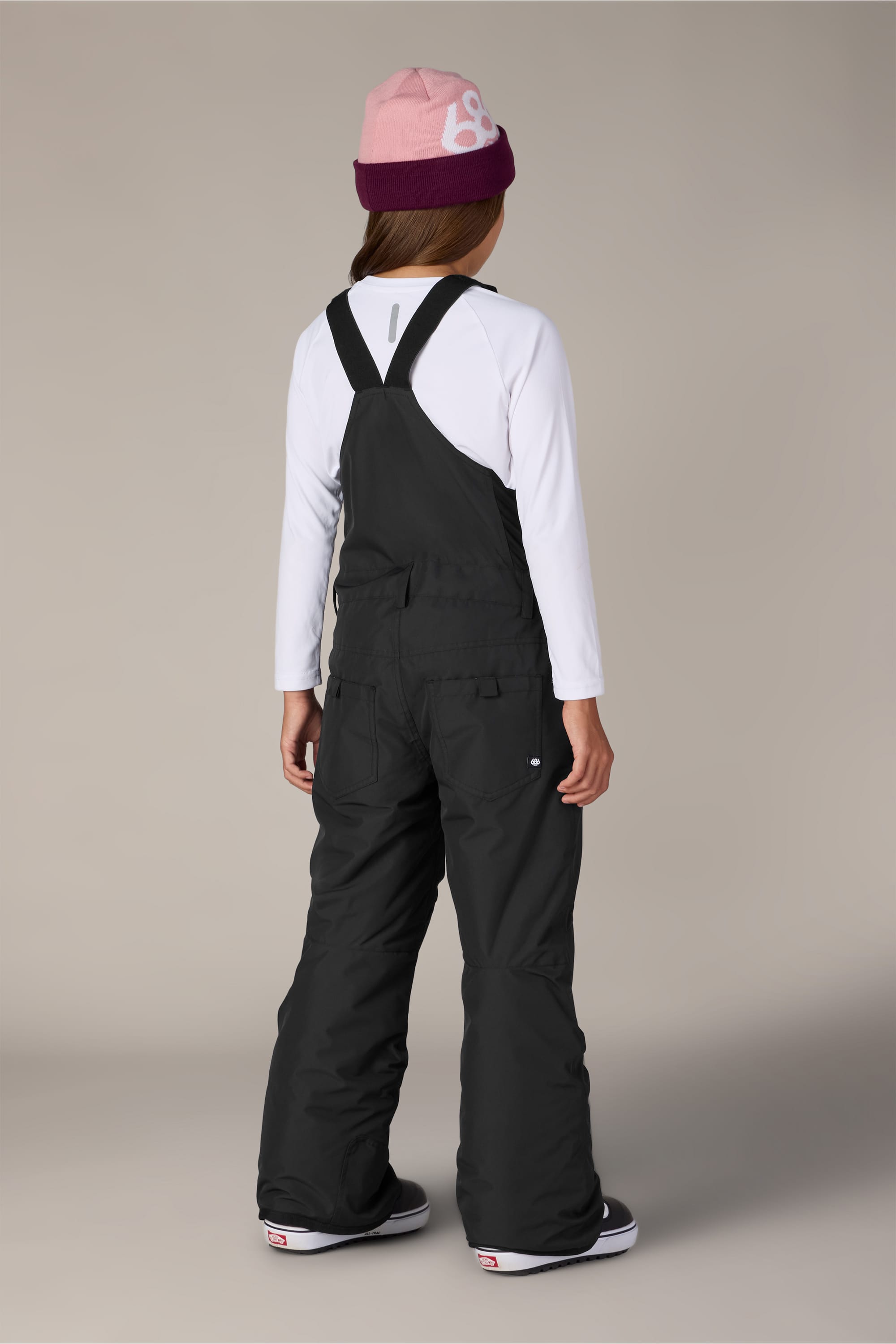 A person stands facing away from the camera against a plain background, wearing a white long-sleeve shirt, the 686 Girls Sierra Insulated Bib by 686 crafted from polyester waterproof material, a pink and burgundy beanie, and sneakers.