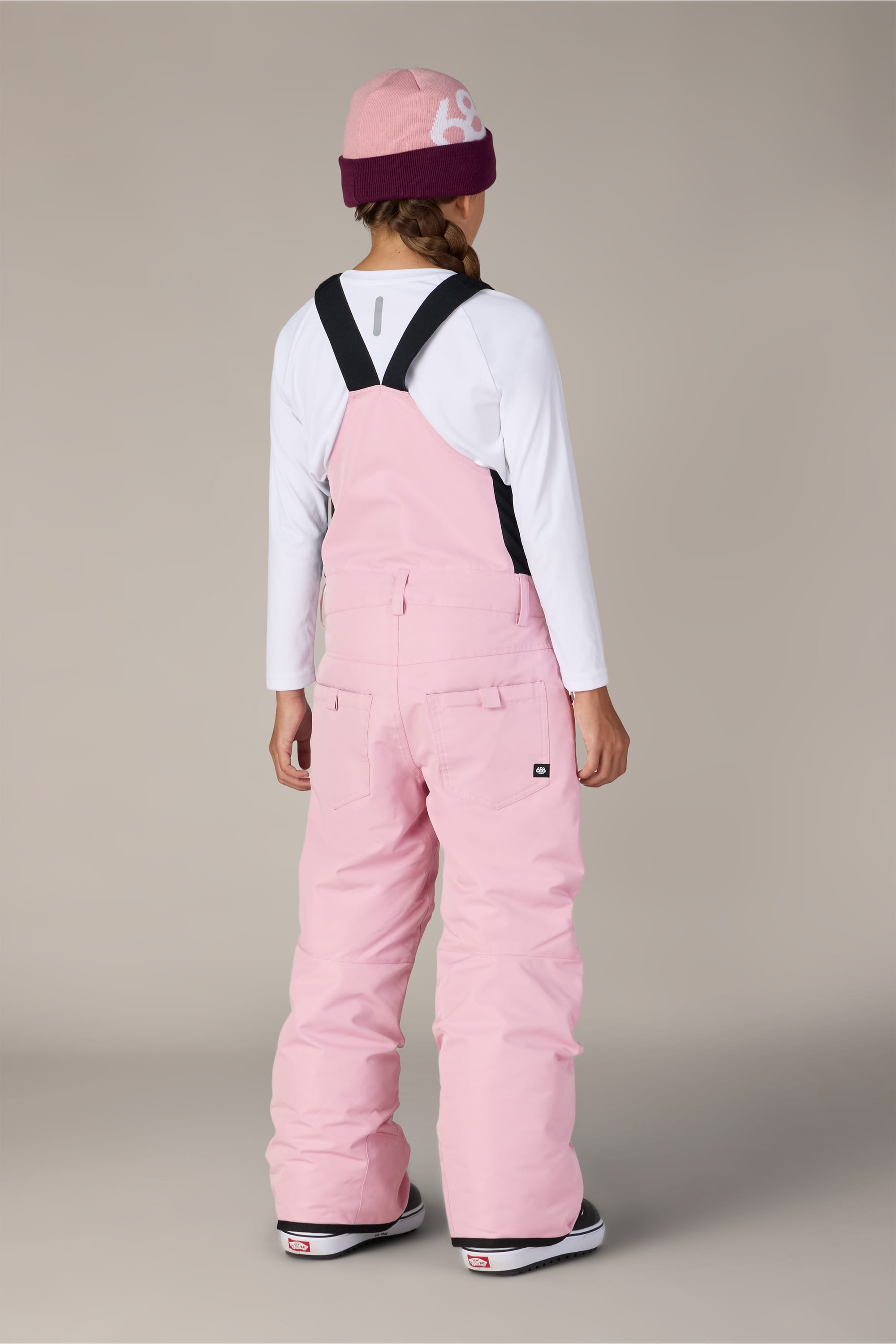 A person stands facing away against a neutral background, wearing a pink beanie, a long-sleeve white shirt, and the 686 Girls Sierra Insulated Bib by 686 in pink.