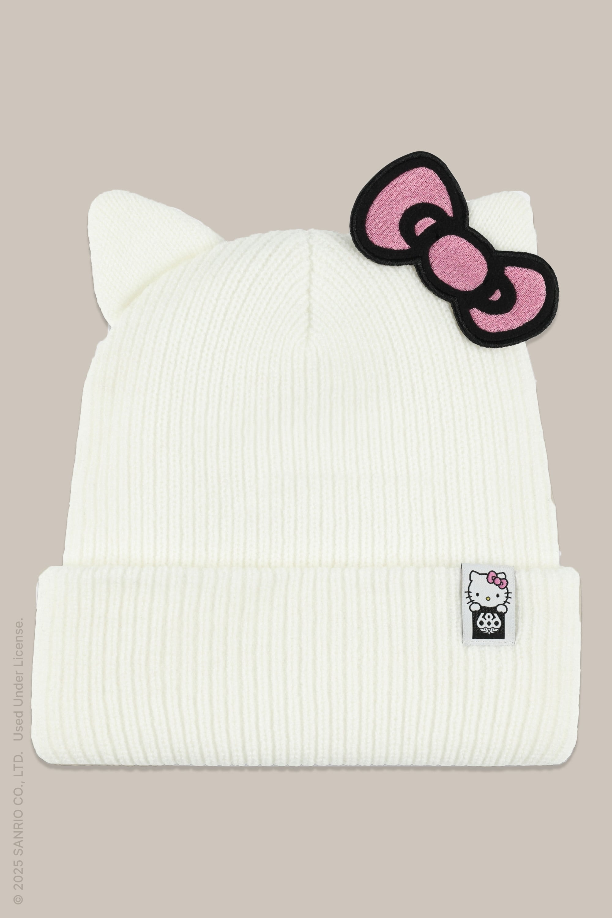 The 686 Women's Hello Kitty® Knit Beanie, in white with cat ears, a pink and black bow on one ear, and a Hello Kitty tag on the front cuff, is shown against a beige background.
