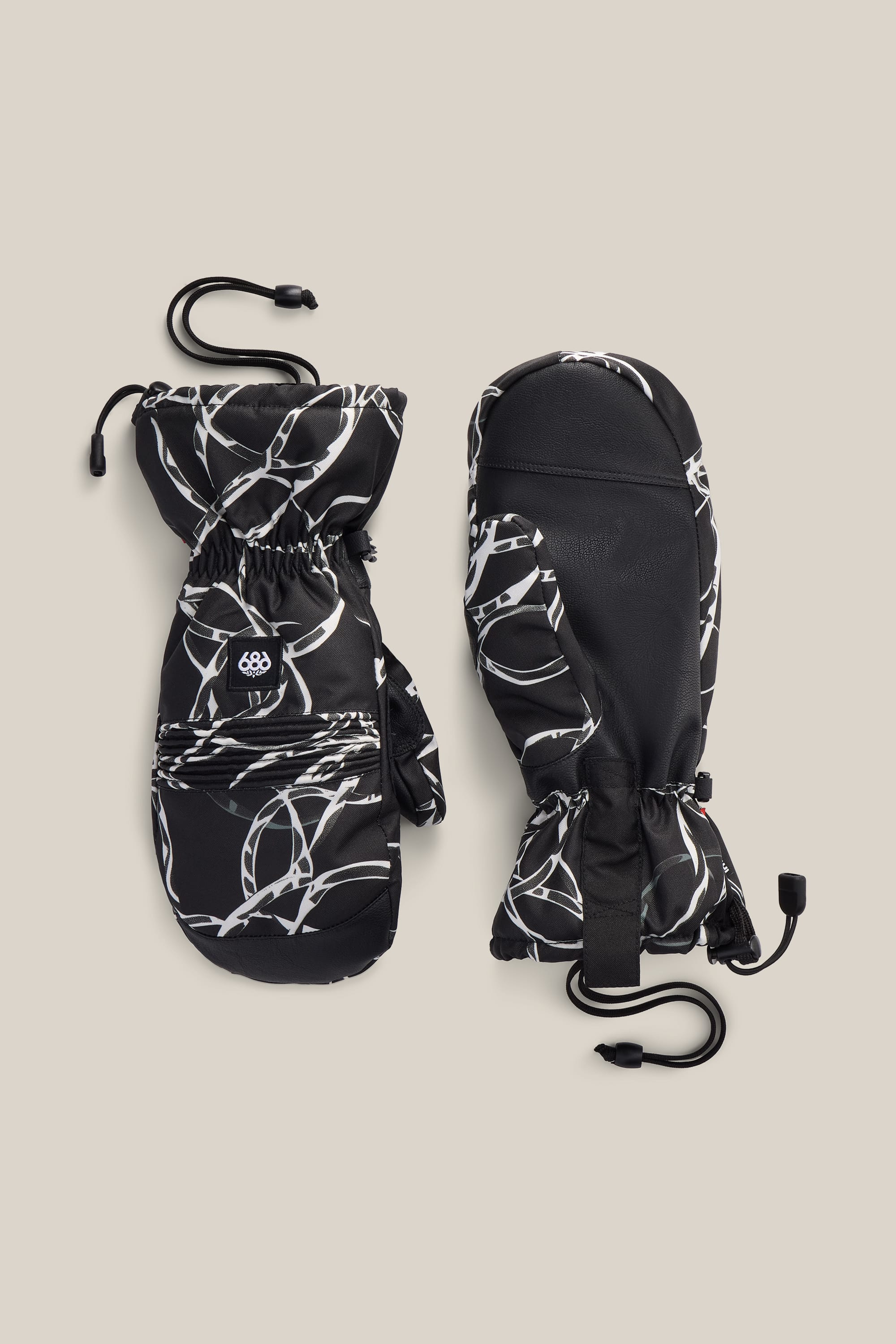A pair of 686 Lander™ Mitts by 686 in black, featuring white abstract line patterns, adjustable wrist straps, and black drawstring cords, displayed on a beige background.