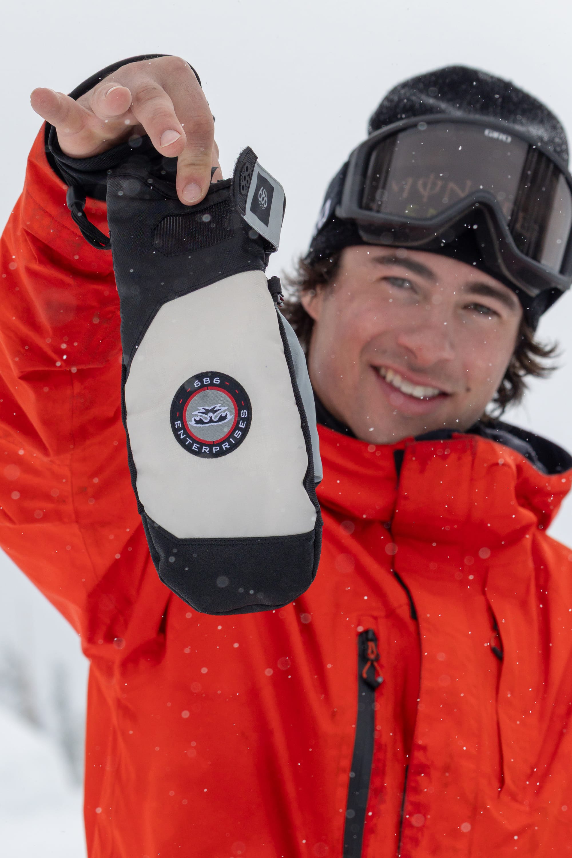 A smiling person in an orange jacket and ski goggles holds up the 686 Men’s Primer Mitt by 686, standing in a snowy outdoor setting.