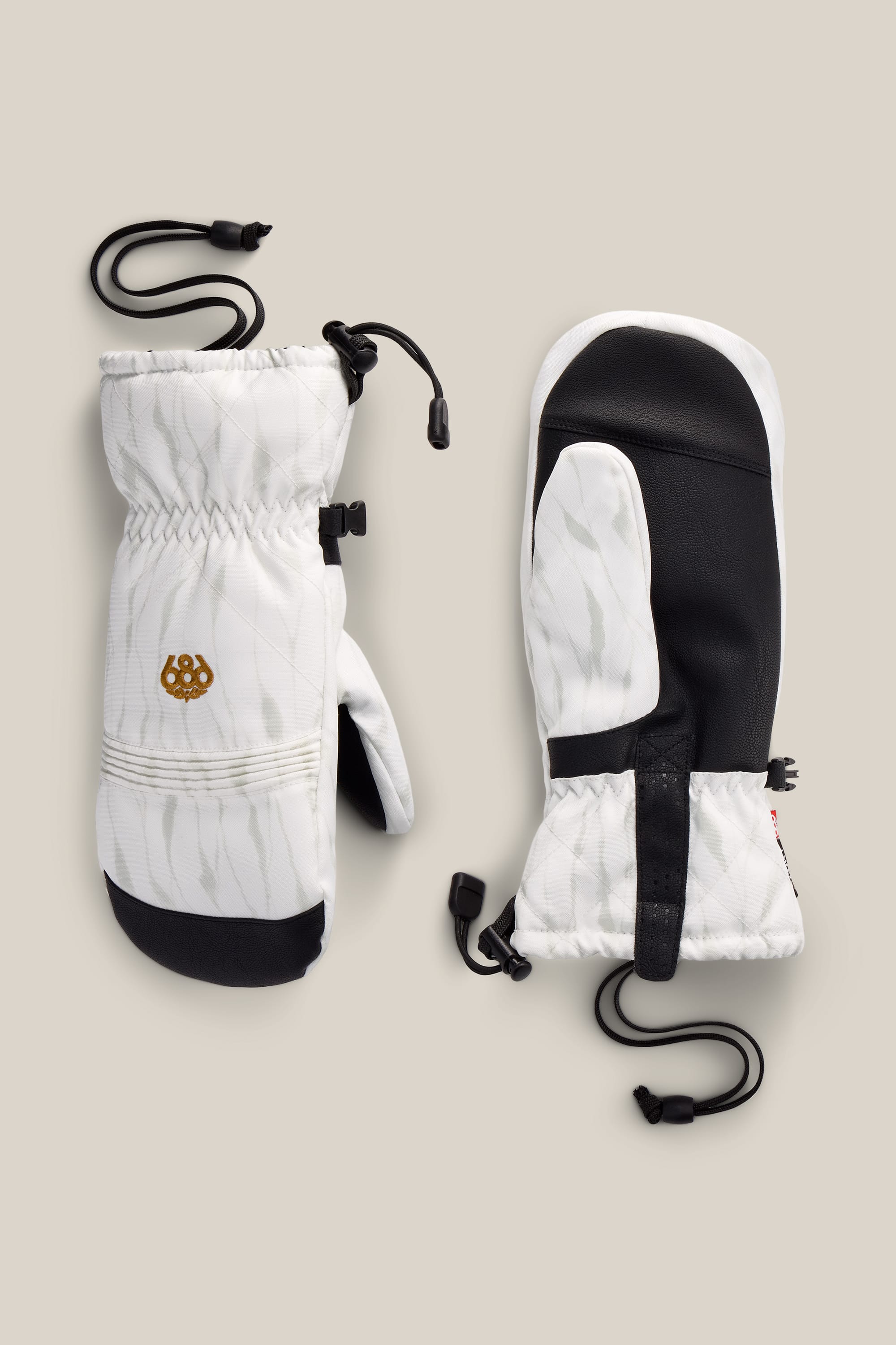 A pair of 686 Women’s Jubilee™ Mitts in white and black, featuring adjustable wrist straps and cords, displayed on a beige background.