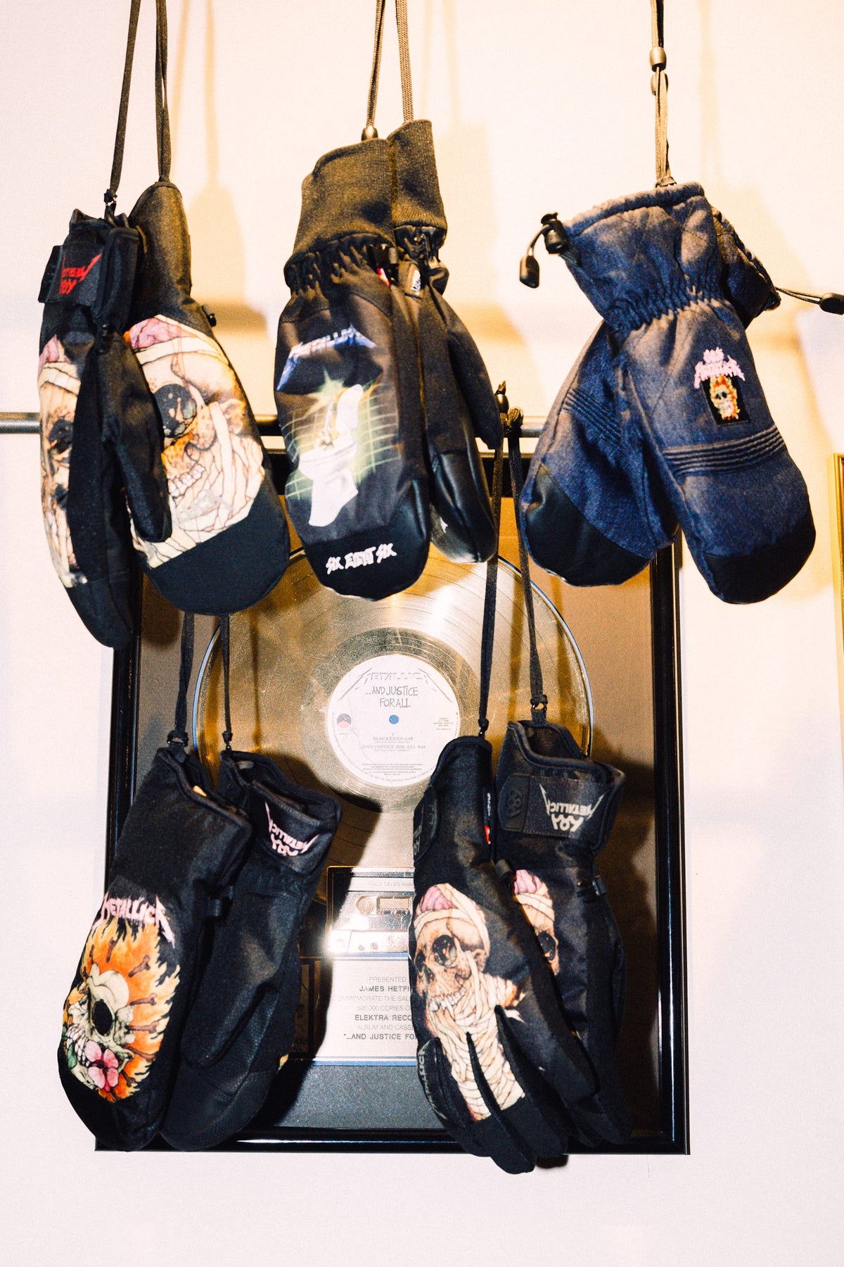 Five pairs of 686 Women’s Revel Mitts by 686, each featuring a unique and colorful design, hang on strings in front of a framed gold record mounted on the wall.