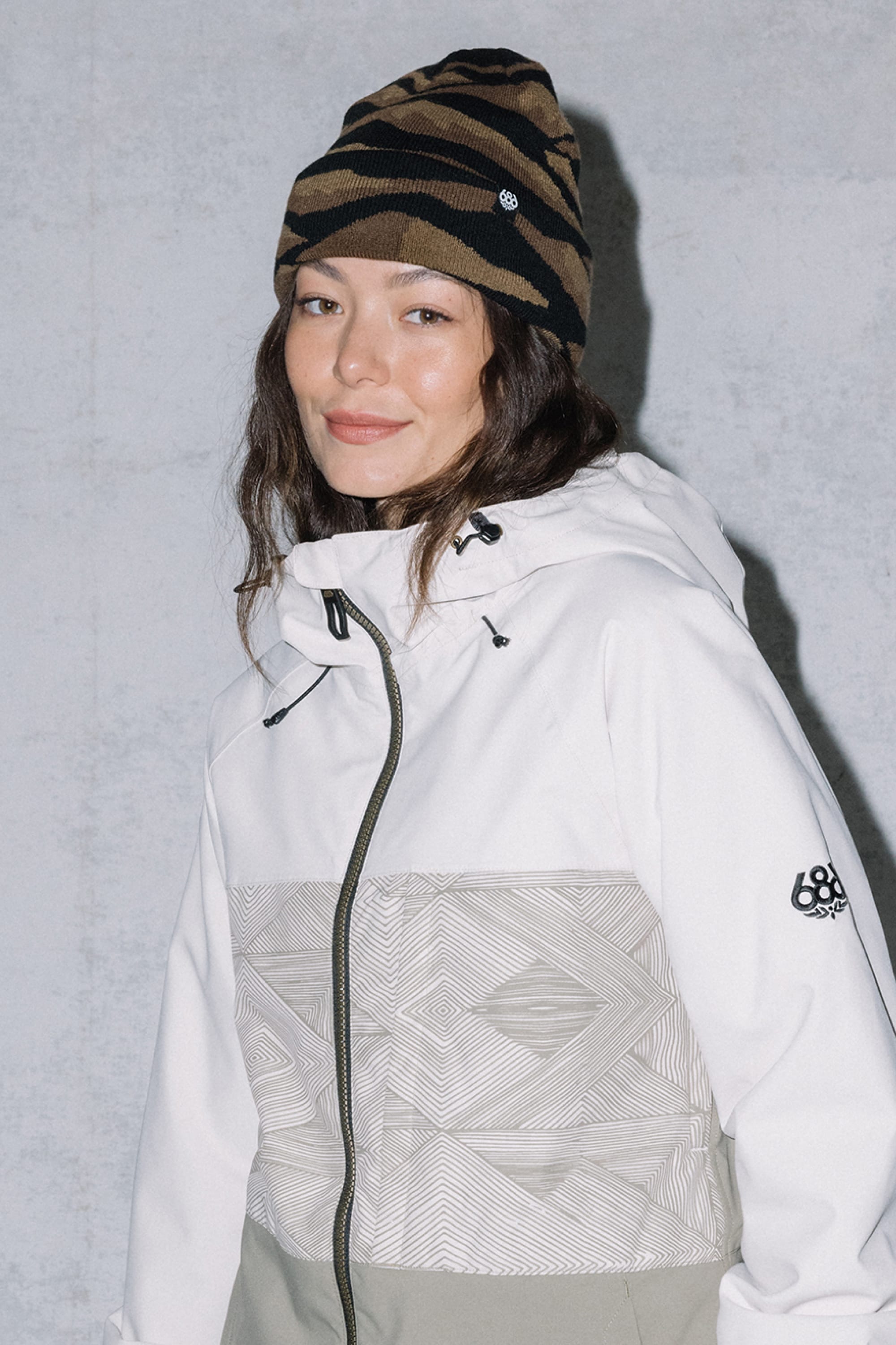 A woman in a white and light gray patterned jacket and the 686 Summit Beanie by 686 stands against a plain light gray wall, smiling at the camera as if recalling epic descents from the highest summit.