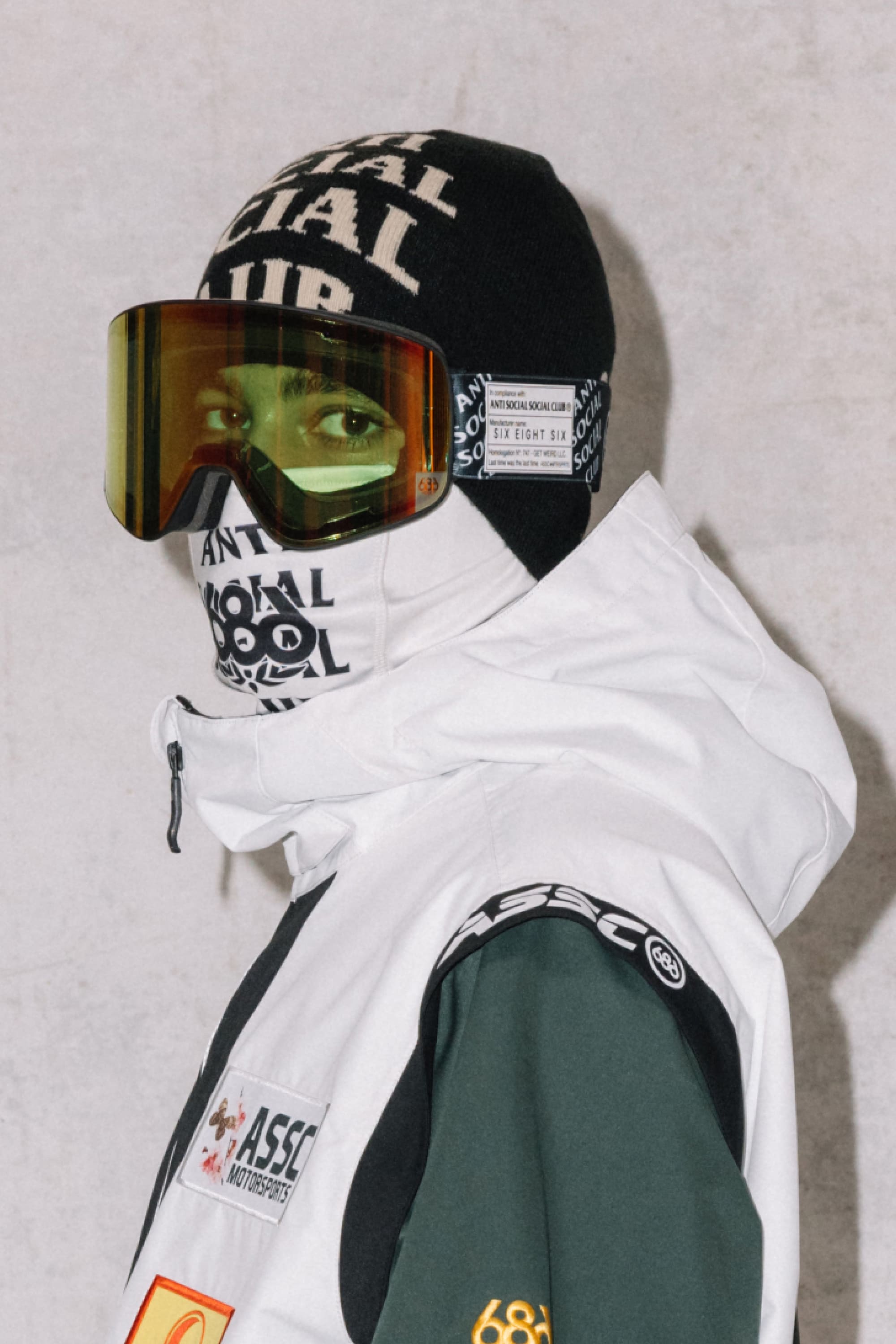 A person in a white and green patched jacket, black beanie, matching face covering with white SOCIAL SOCIAL CLUB text, and large 686 SMARTY® Magnetic Goggles by 686 stands against a light background.