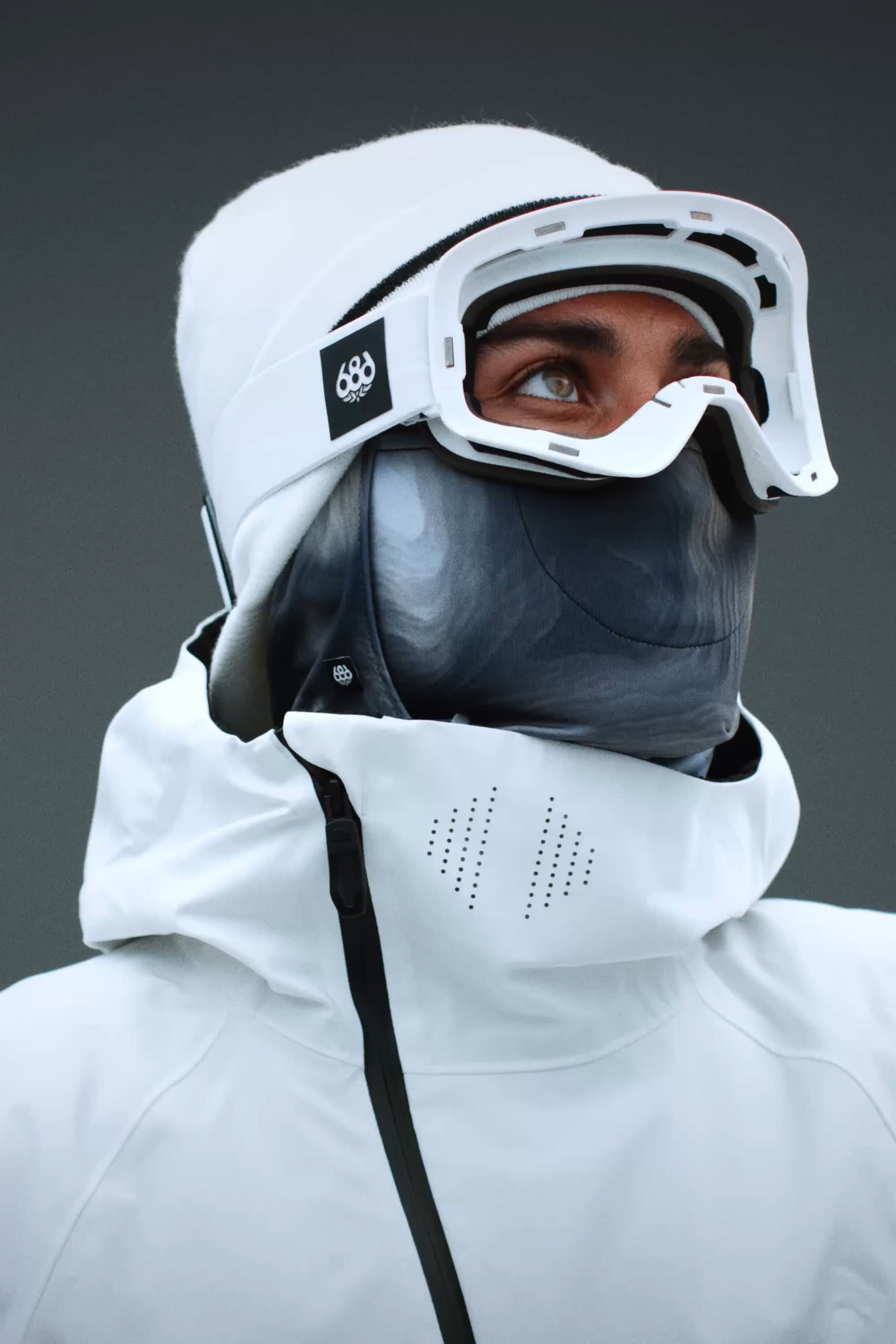 A person wearing a white jacket with a high collar, a white beanie, gray face mask, and 686 SMARTY® Magnetic Goggle by 686 looks upward against a plain dark background.