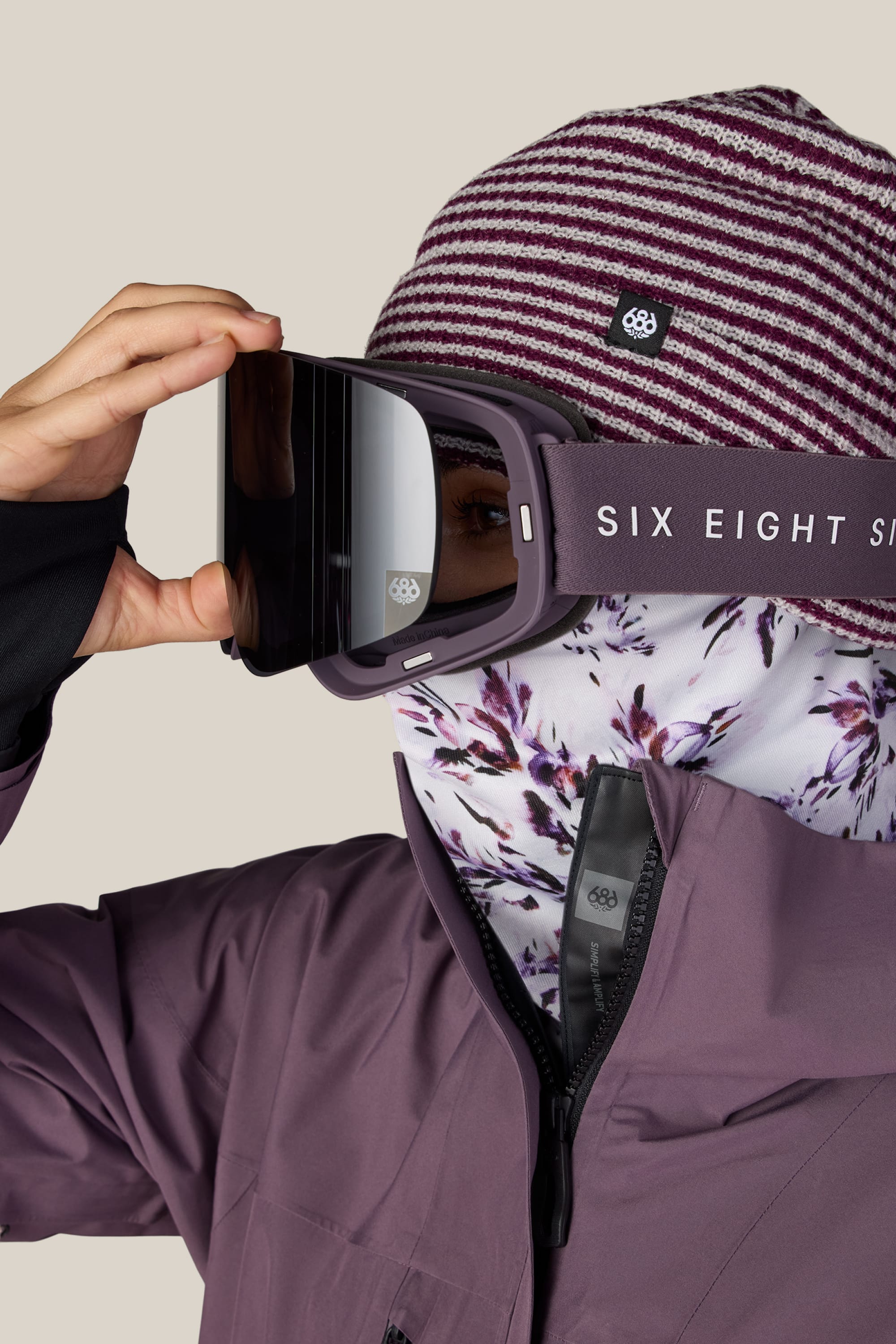 A person in a striped beanie, floral face covering, and ski jacket adjusts reflective 686 SMARTY® Magnetic Goggles featuring the 686 logo on the strap.