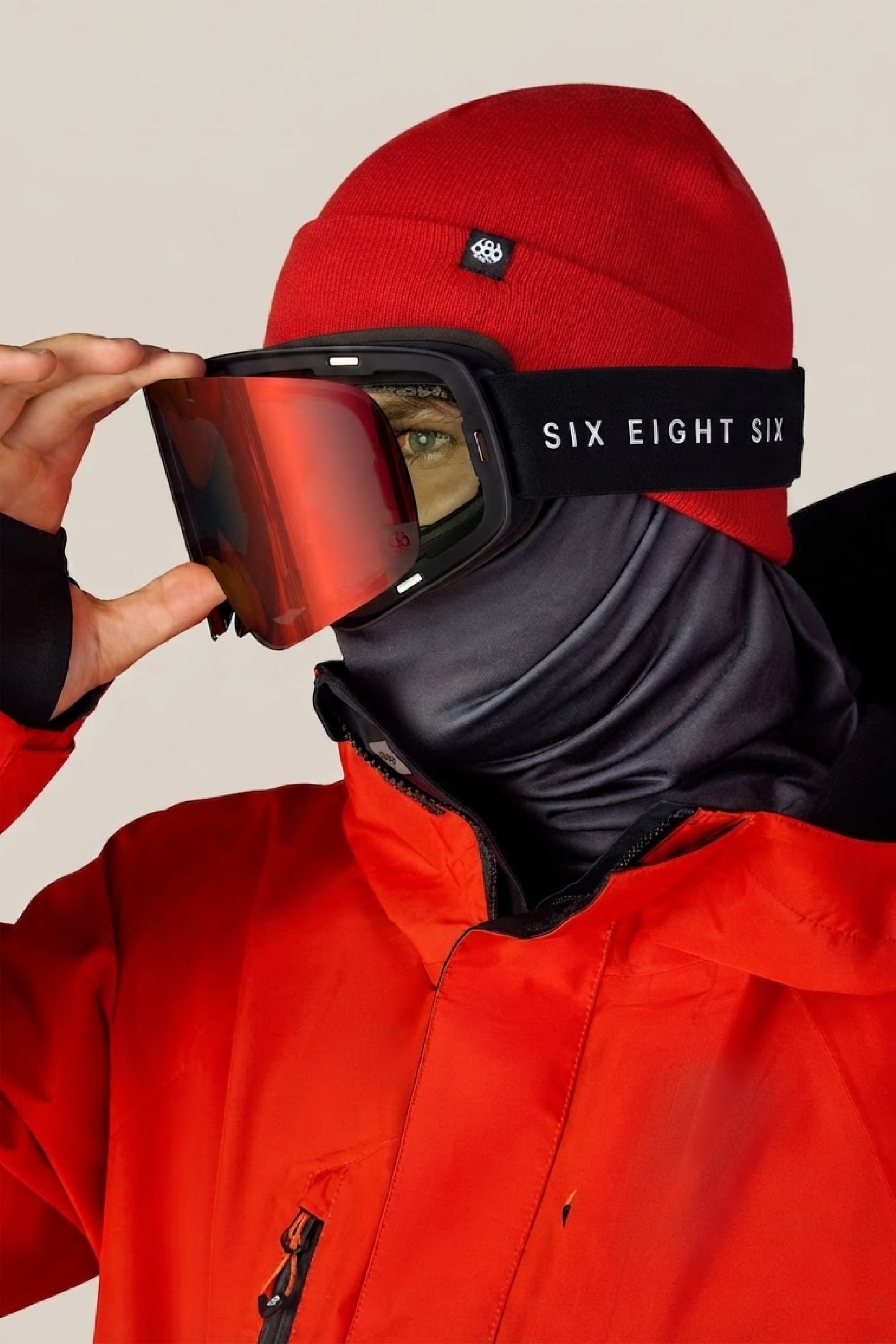 A person in a red jacket and beanie adjusts 686 SMARTY® Magnetic Goggles, featuring a 686 strap, while wearing a black face covering.