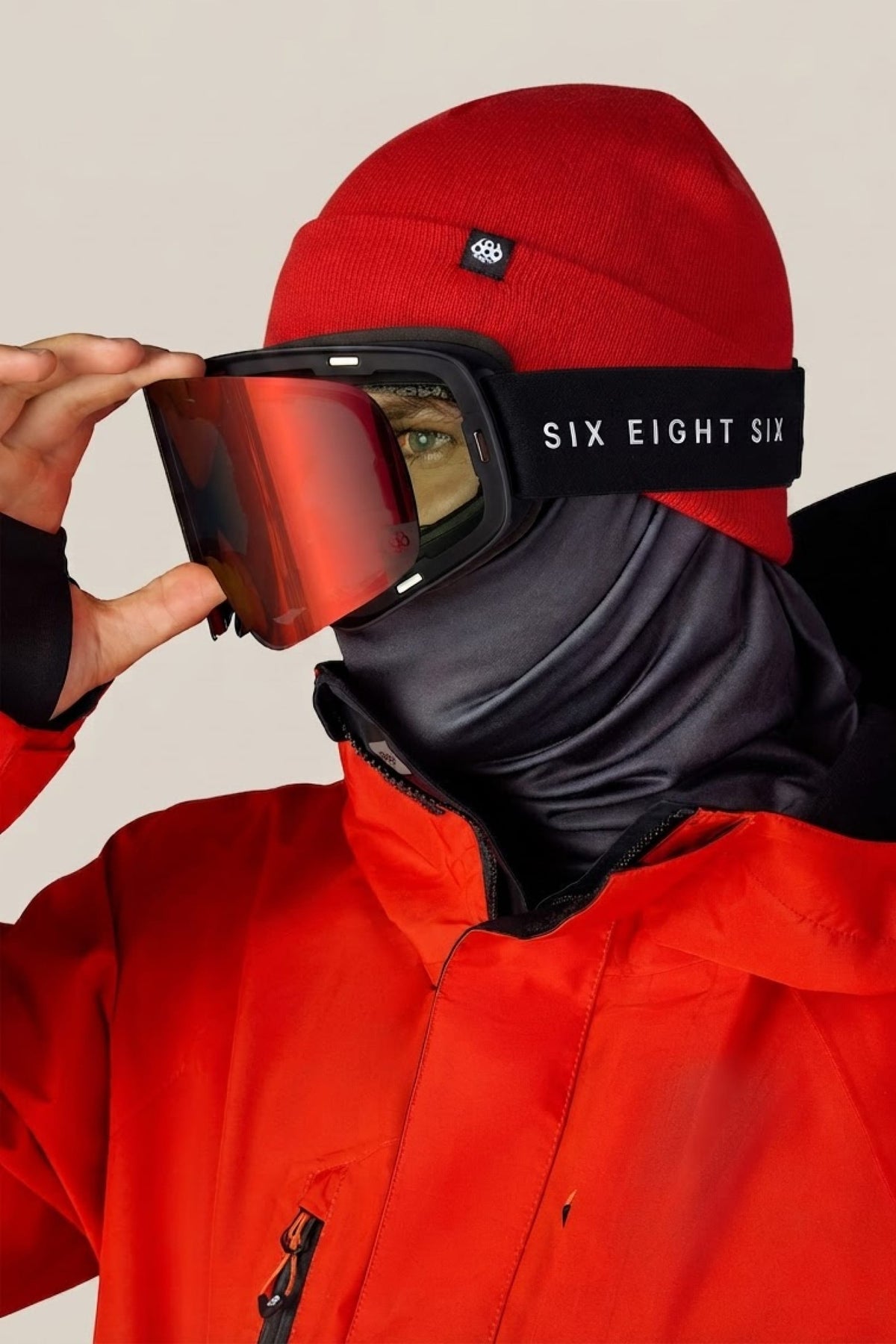 A person in a red jacket and beanie adjusts their 686 SMARTY® Magnetic Goggle, featuring a black face covering and visible 686 branding on the goggle strap.