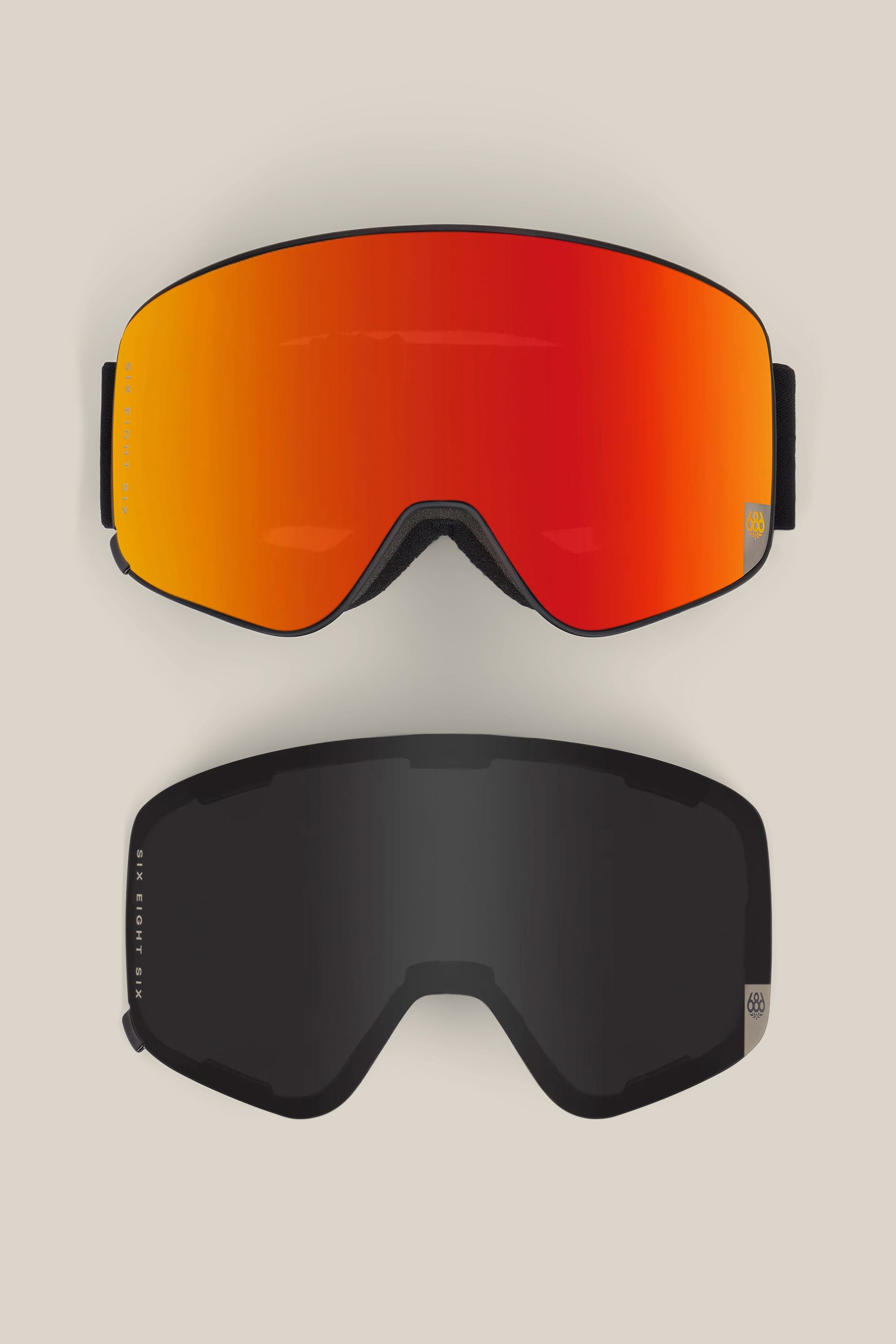 Two 686 SMARTY® Magnetic Goggles by 686, each featuring interchangeable lenses: the top goggle has a red-orange reflective lens, while the bottom goggle has a dark non-reflective lens. Both have black frames with a small round logo on the bottom right corner.