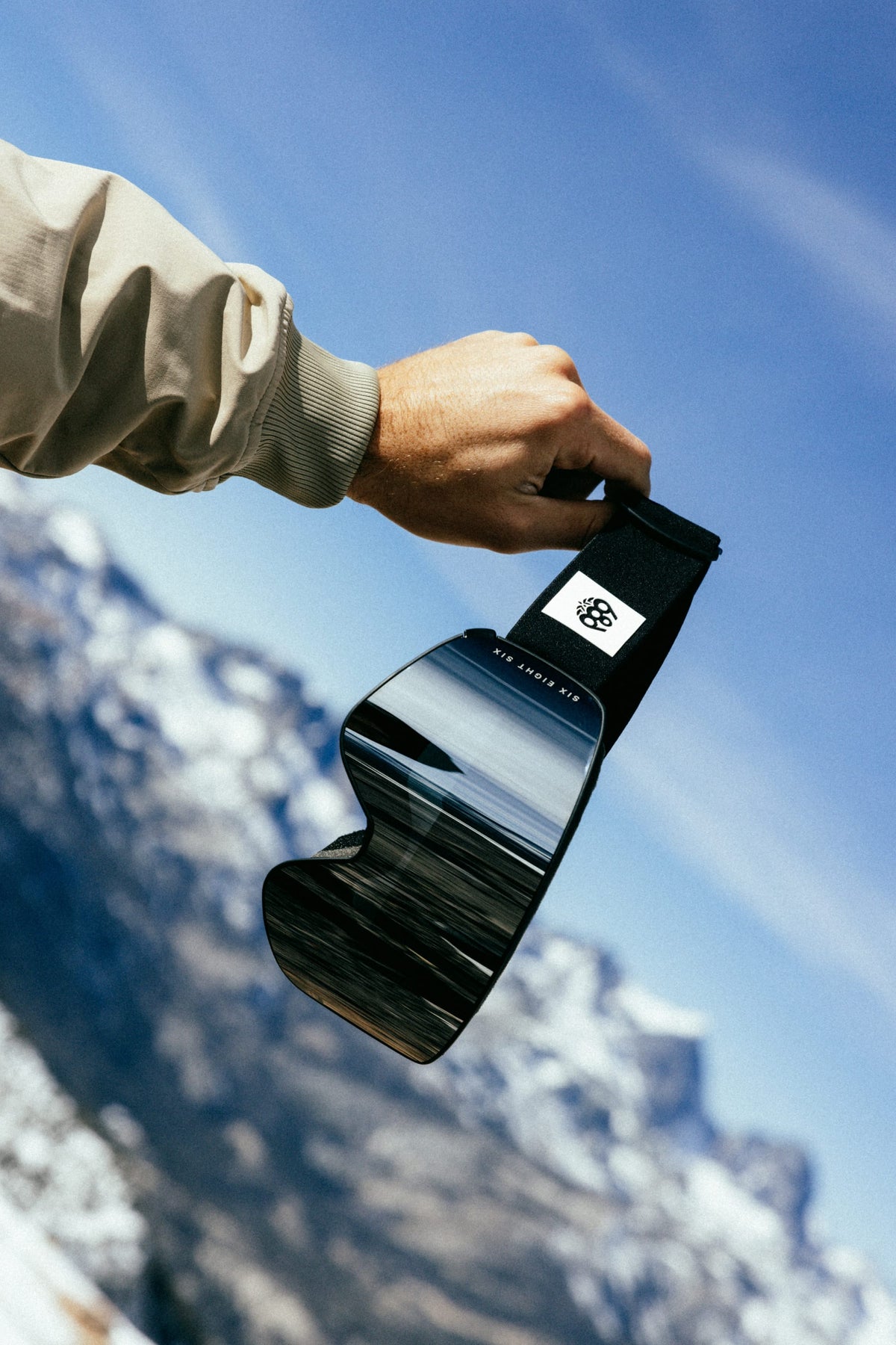 A hand holds the 686 SMARTY® Magnetic Goggle by 686, featuring a black strap and a reflective lens that displays a mountain landscape in its reflection. Snowy mountains and a blue sky can be seen in the background.
