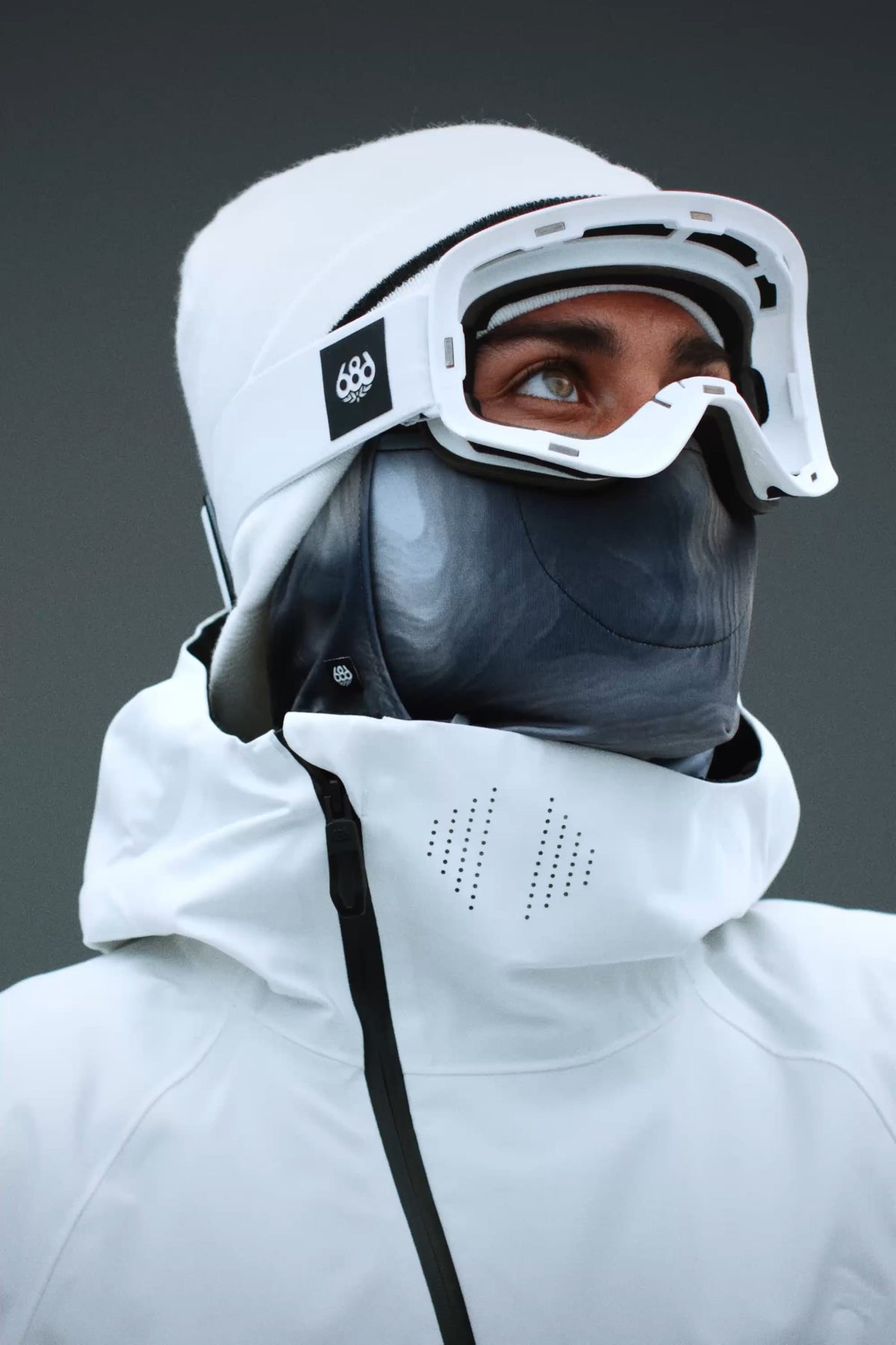 A person wearing the 686 SMARTY® Magnetic Goggle by 686, along with a white jacket, white hat, and dark face mask, looks upward against a plain gray background.