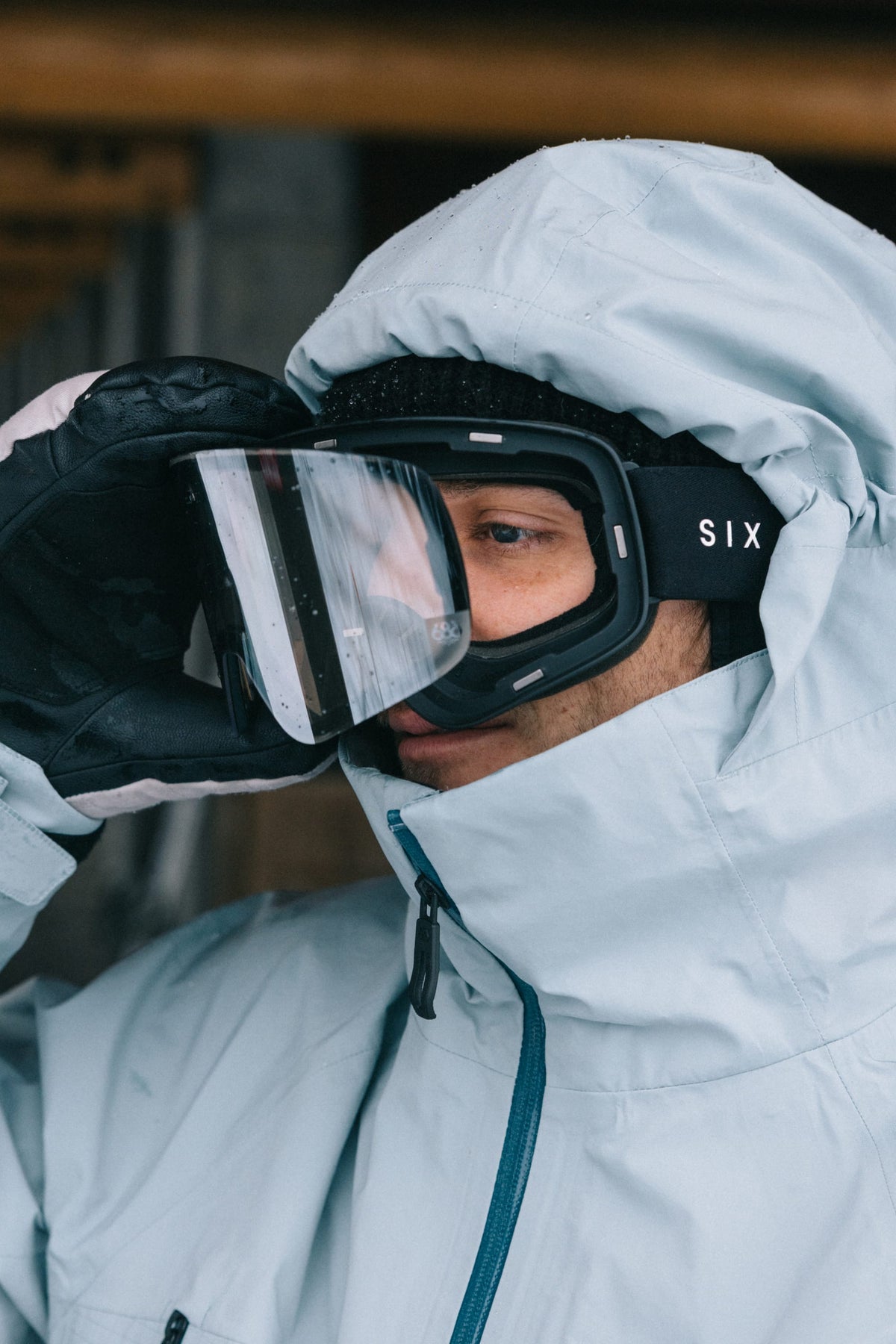 A person in a light blue winter jacket and black 686 SMARTY® Magnetic Goggle by 686 looks into the distance, adjusting the goggles with a gloved hand.
