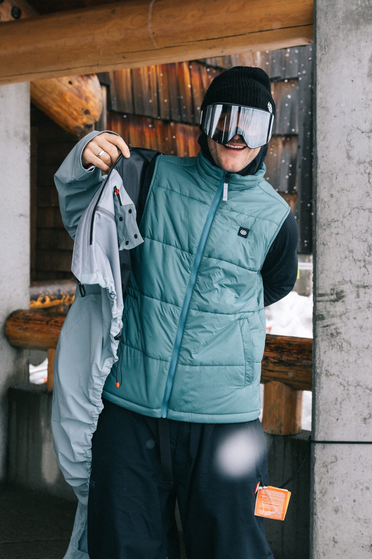 A person wearing a teal puffer vest, black knit hat, and 686 SMARTY® Magnetic Goggle by 686 stands under a rustic shelter, holding a gray jacket with snow visible in the background.